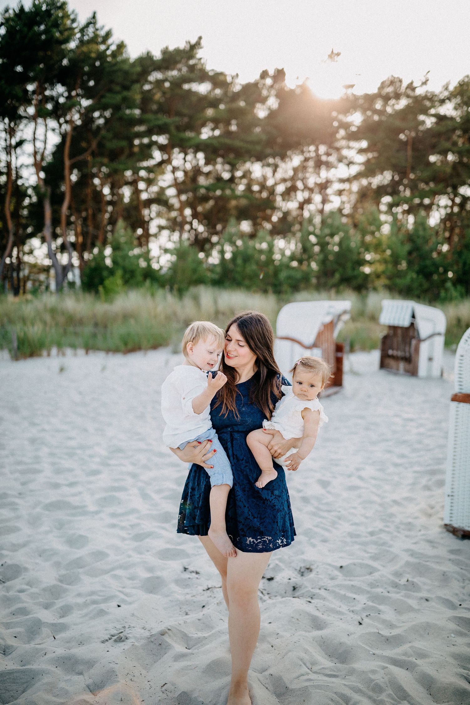 Mutter in marineblauem Kleid hält zwei Kinder am sonnigen Strand mit Liegestühlen im Hintergrund.