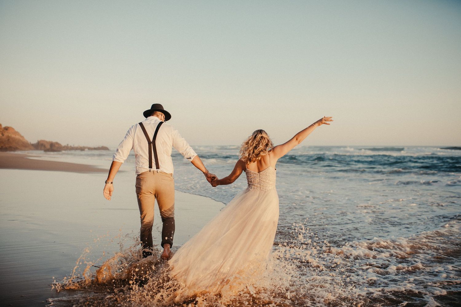 Ein Paar in Hochzeitskleidung läuft spielerisch durch die Meereswellen an einem Strand bei Sonnenuntergang.