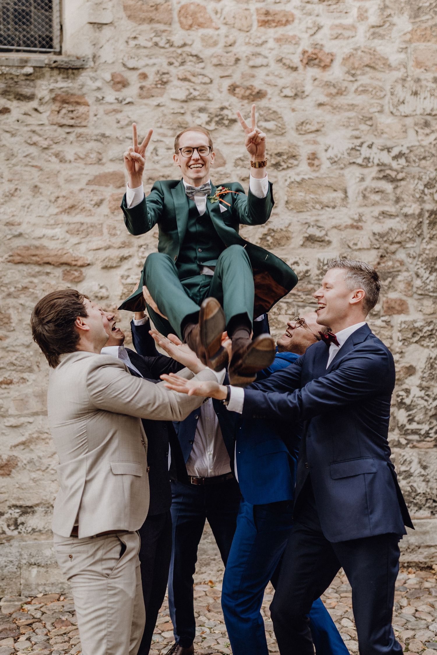 Group of groomsmen in suits lifting and celebrating at a wedding venue against rustic stone wall.