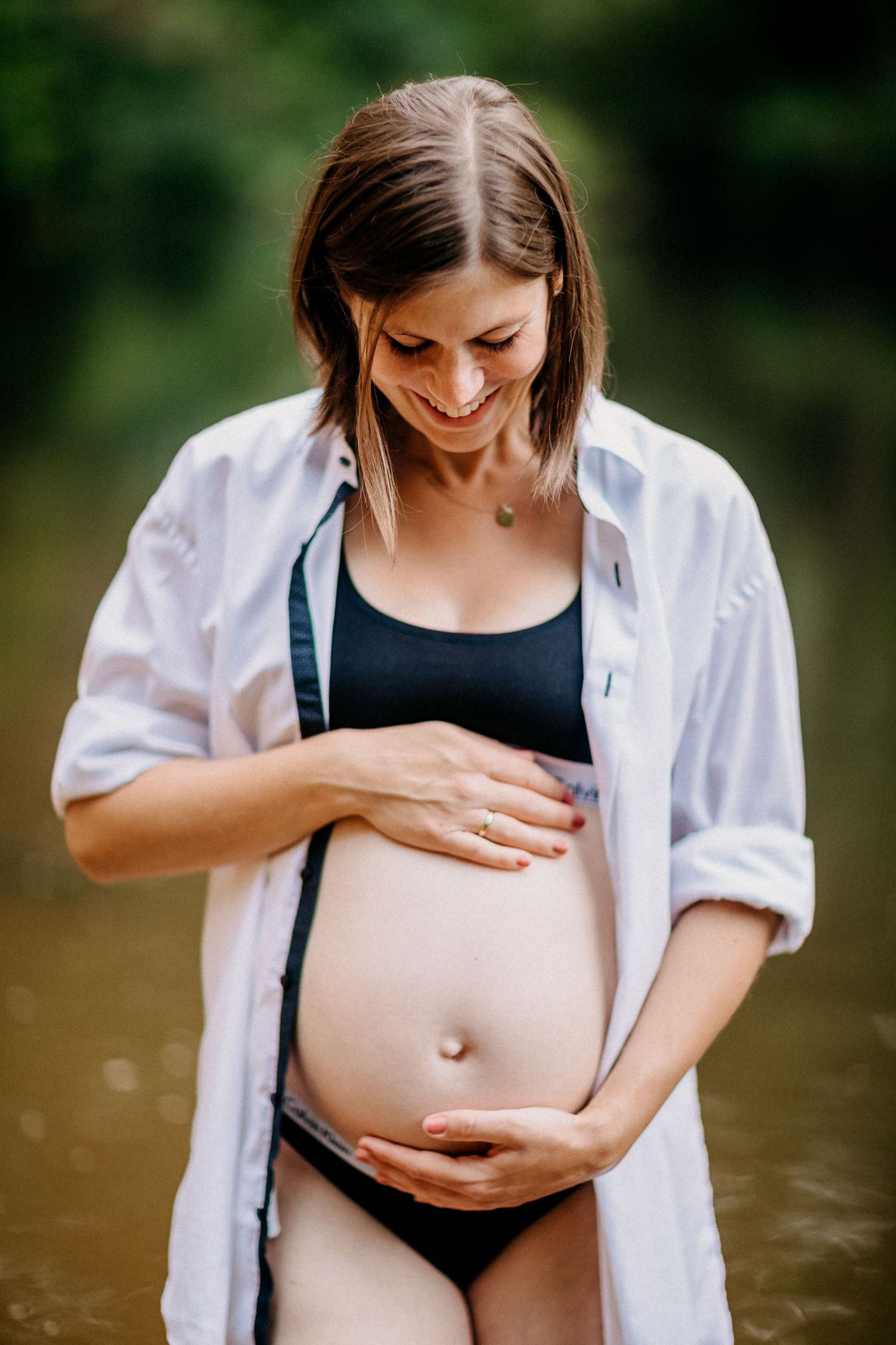 Werdende Mutter in weißem Hemd und Badebekleidung berührt ihren Babybauch am Wasser.