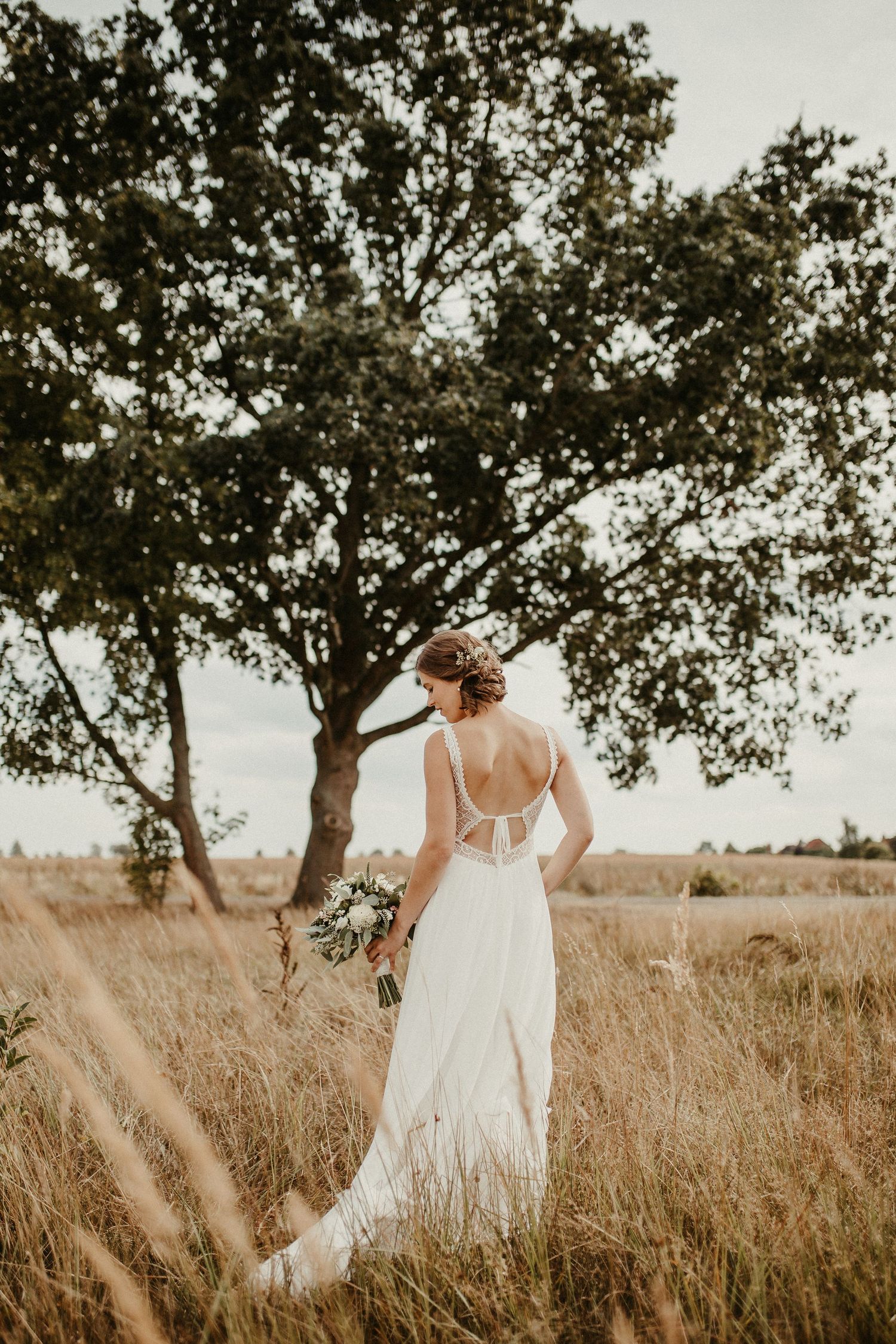 Braut im rückenfreien Hochzeitskleid, das Blumenstrauß hält, während sie bei Sonnenuntergang auf goldenem Feld steht.