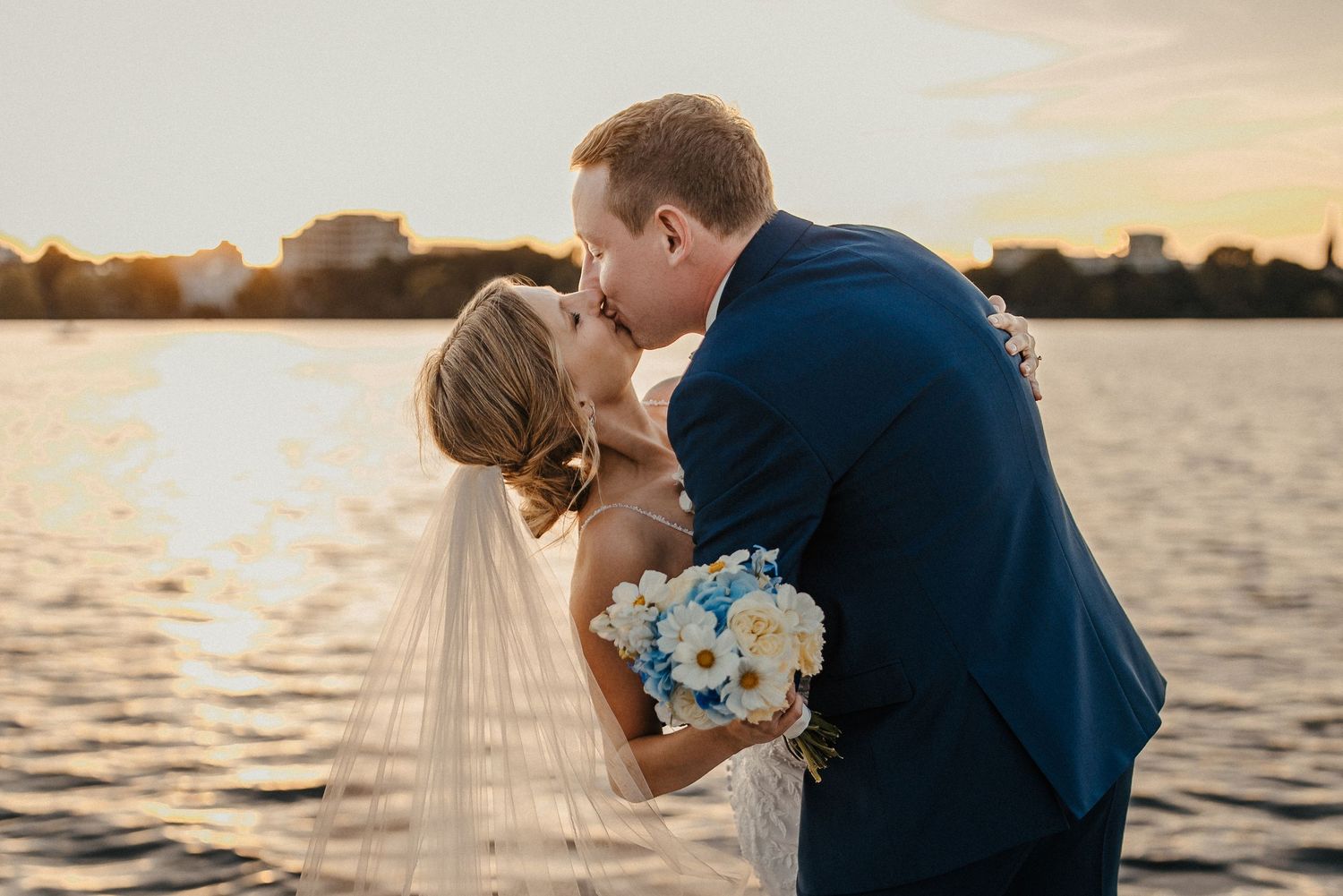 Sonnenuntergangsporträt am Wasser eines Paares, das einen intimen Kuss mit blauem Blumenstrauß teilt in Hamburg an der Alster.