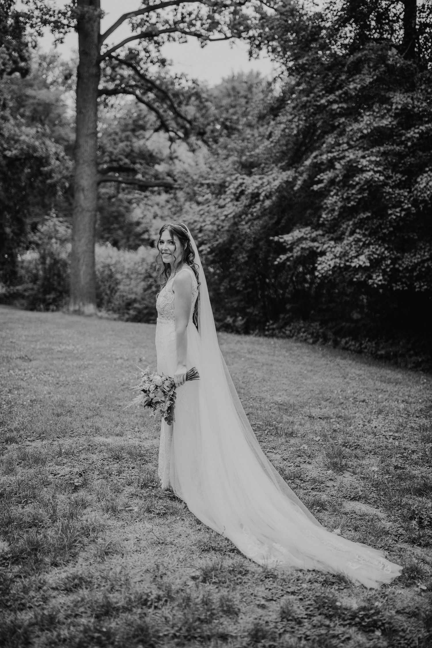 Eine Braut in einem fließenden weißen Kleid und Schleier steht in einem Garten und hält einen Blumenstrauß in Schwarz-Weiß-Fotografie.