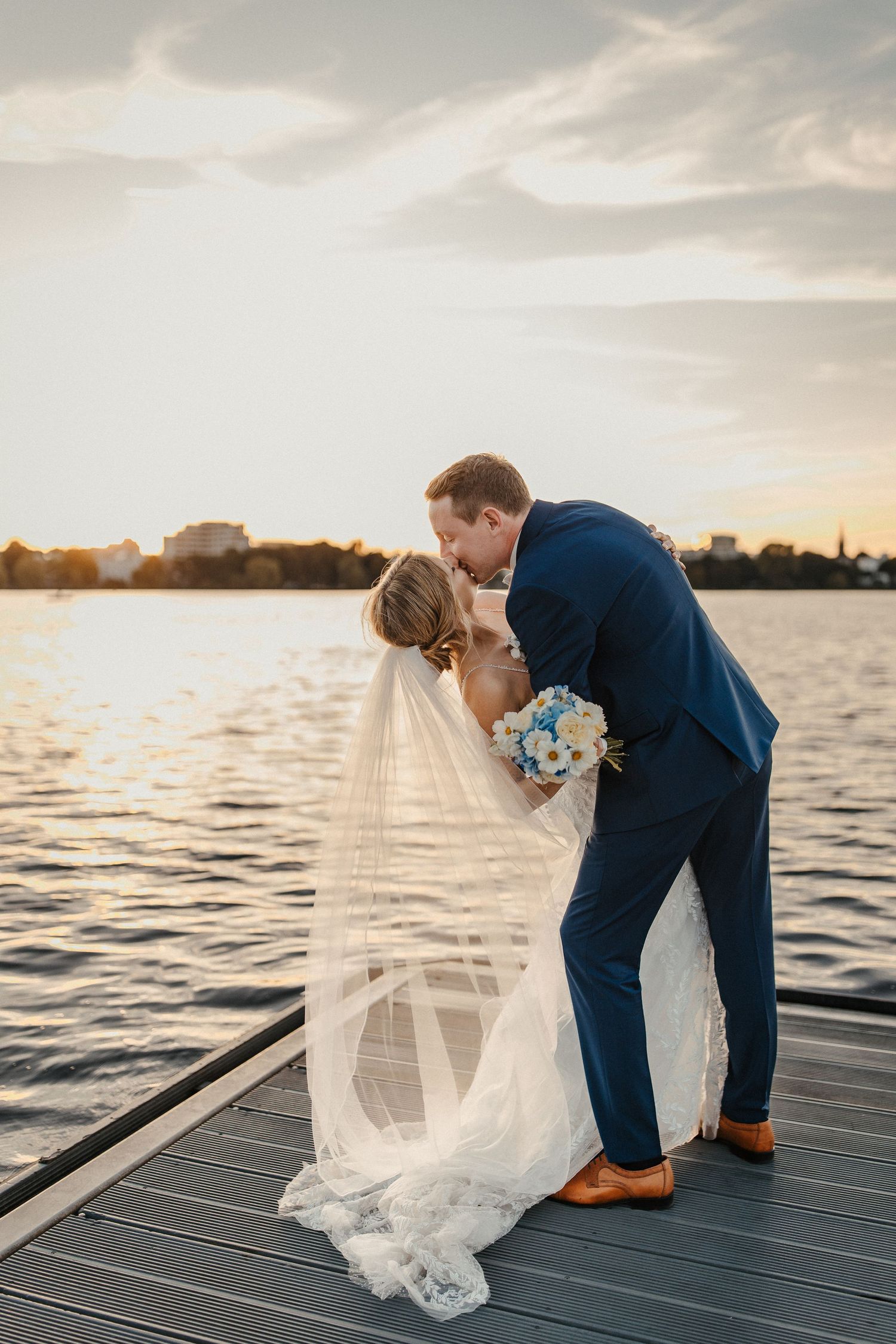 Romantischer Sonnenuntergangskuss zwischen Jungvermählten am Wasserdock mit fließendem Hochzeitskleid in Hamburg an der Alster.