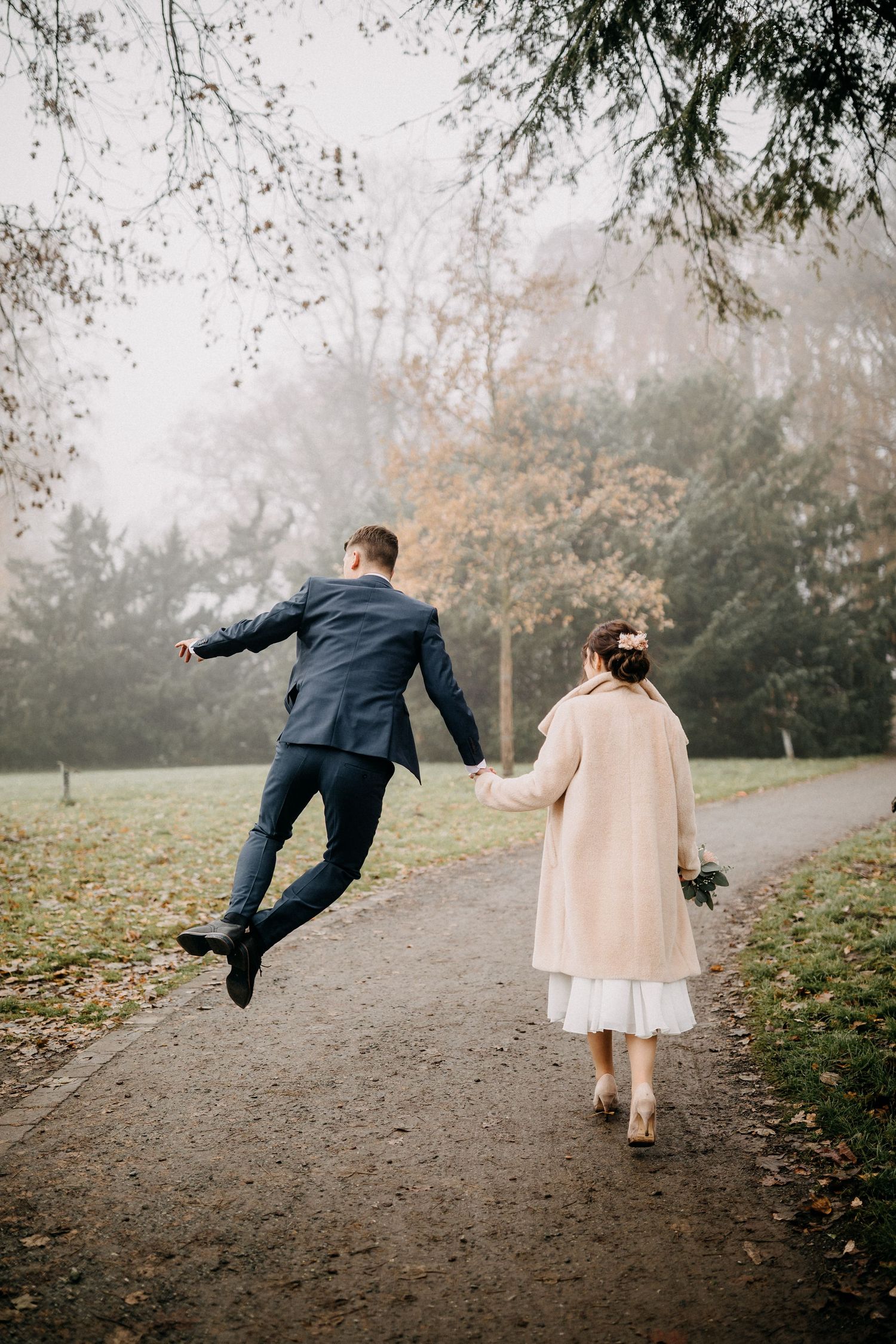 Verspieltes Hochzeitsfoto eines Paares auf dem Weg mit einer Person, die in die Luft springt in Wolfenbüttel.