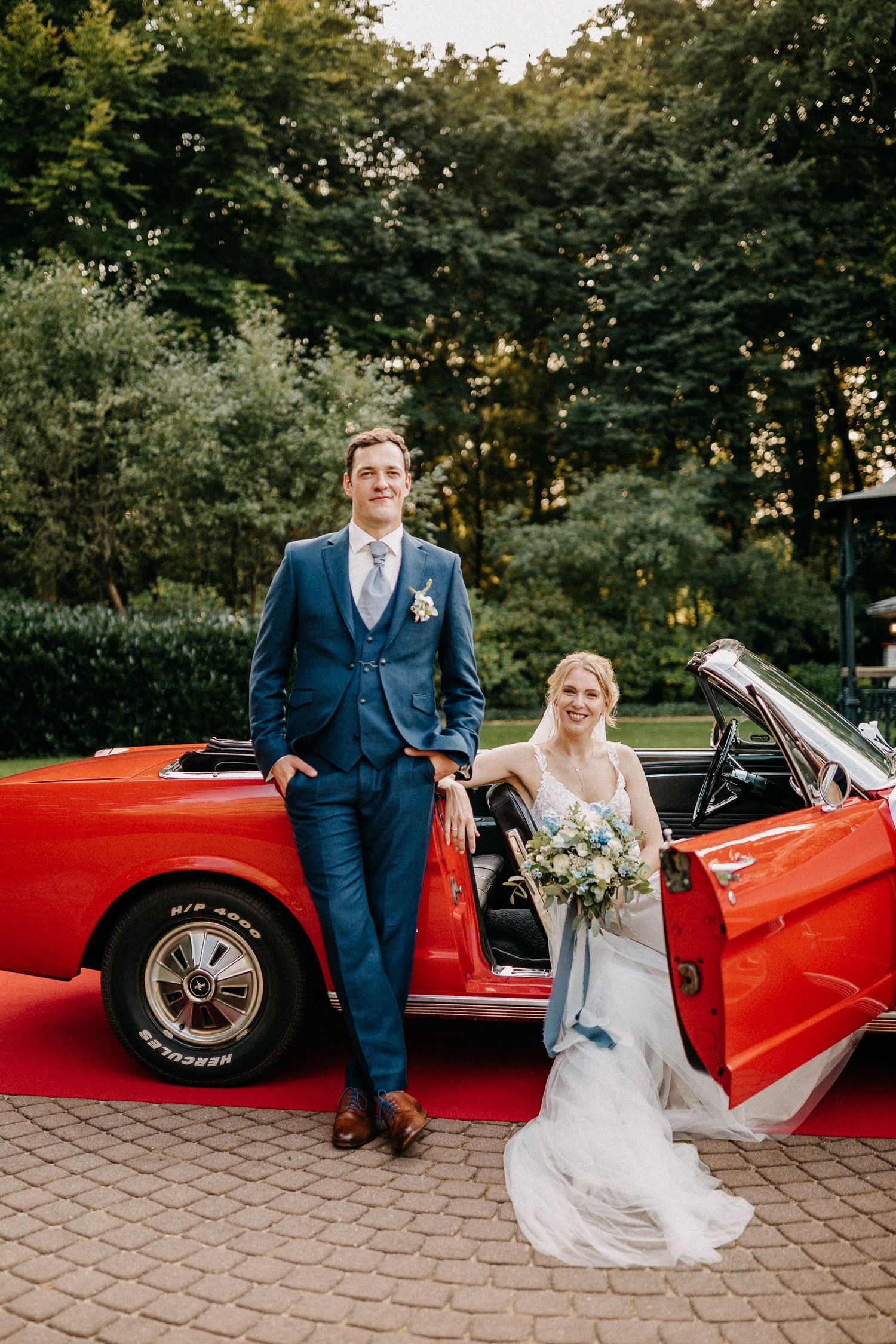 Frisch verheiratetes Paar posiert mit rotem Cabrio-Oldity am Veranstaltungsort im Freien.
