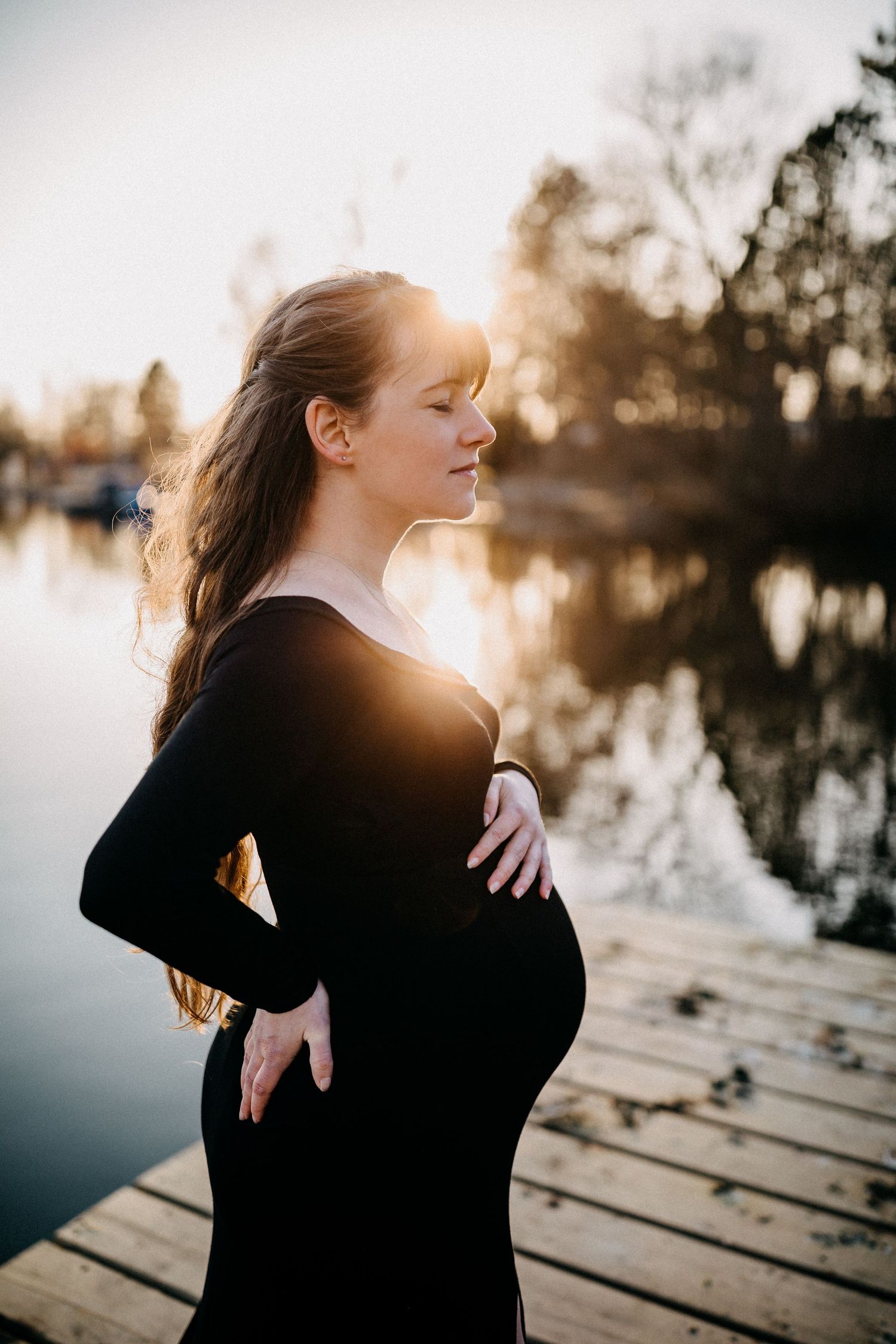 Silhouette einer schwangeren Frau im schwarzen Kleid, die bei Sonnenuntergang auf dem Dock steht.