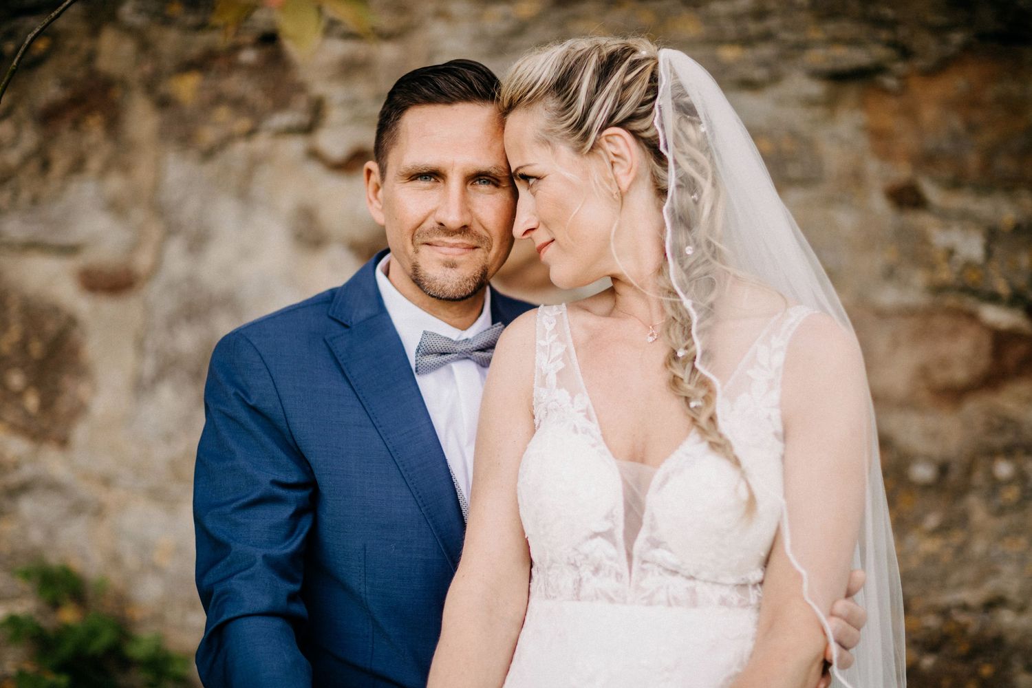 Ein intimes Hochzeitsporträt eines Paares in einem marineblauen Anzug und einem weißen Kleid gegen eine Steinmauer.