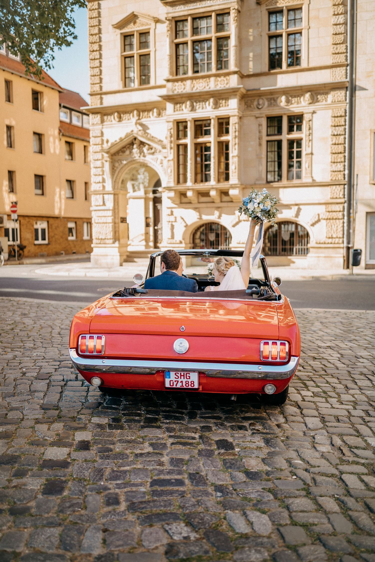 Ein klassisches rotes Ford Mustang Cabrio vor einem historischen europäischen Gebäude auf Kopfsteinpflasterstraßen geparkt.