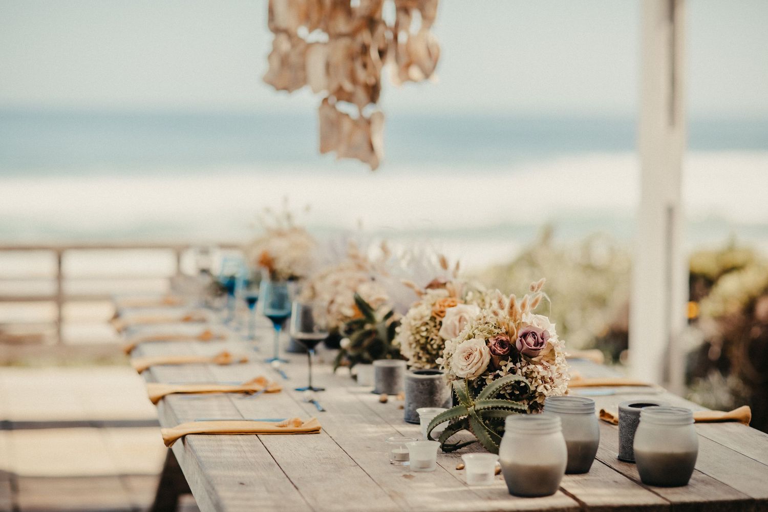 Eleganter Hochzeitsempfangstisch am Strand mit floralen Mittelstücken und Meerblick.