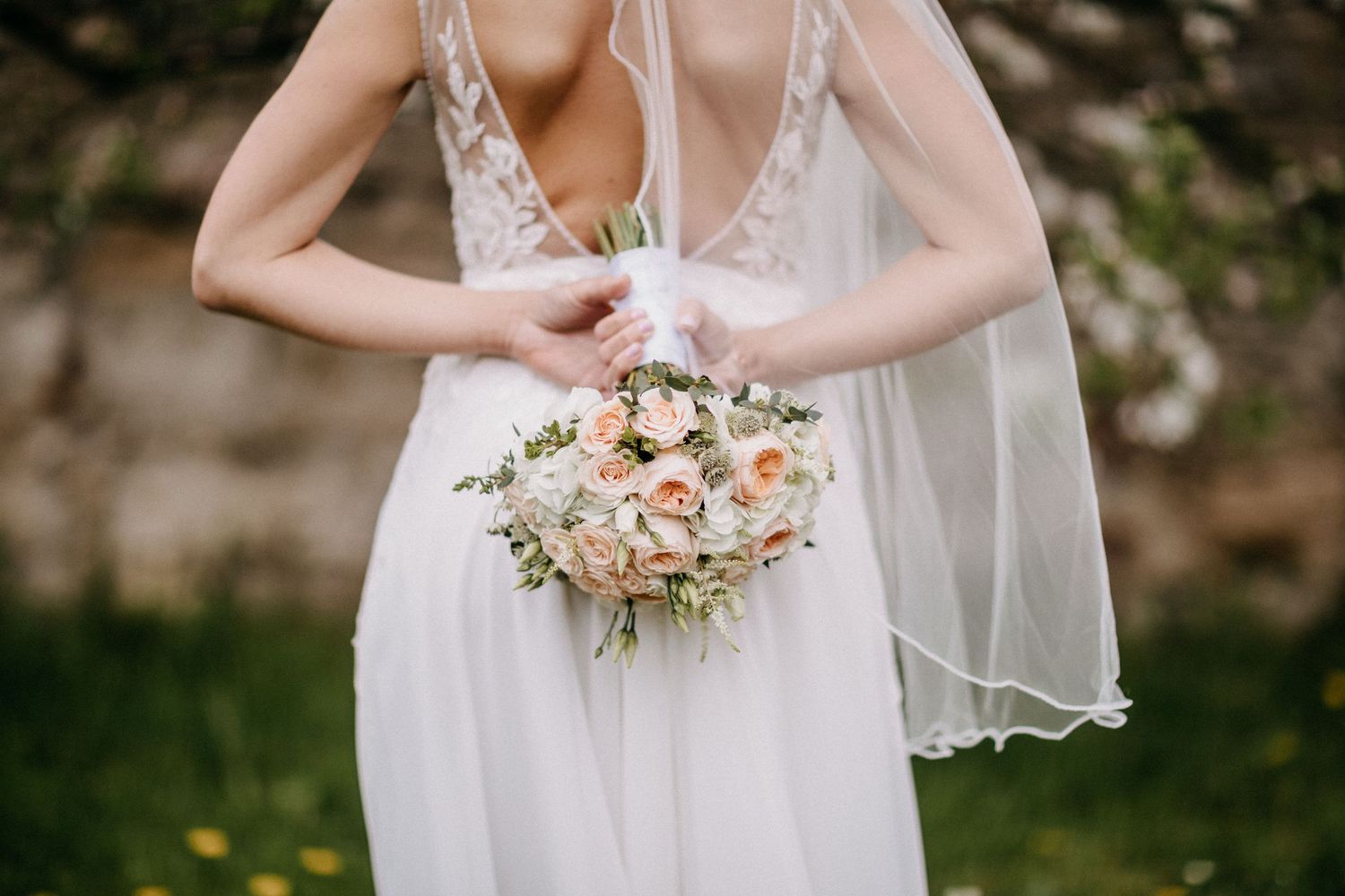 Nahaufnahme des zarten Brautkleides aus Spitze der Braut und des rosa Blumenstraußes von hinten.