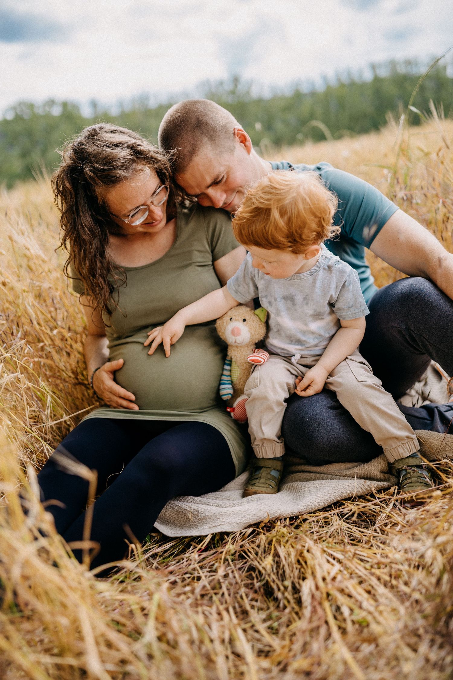 Familie sitzt zusammen auf einem hohen Grasfeld mit Kleinkind, das Stofftier hält und Babybauch streichelt.