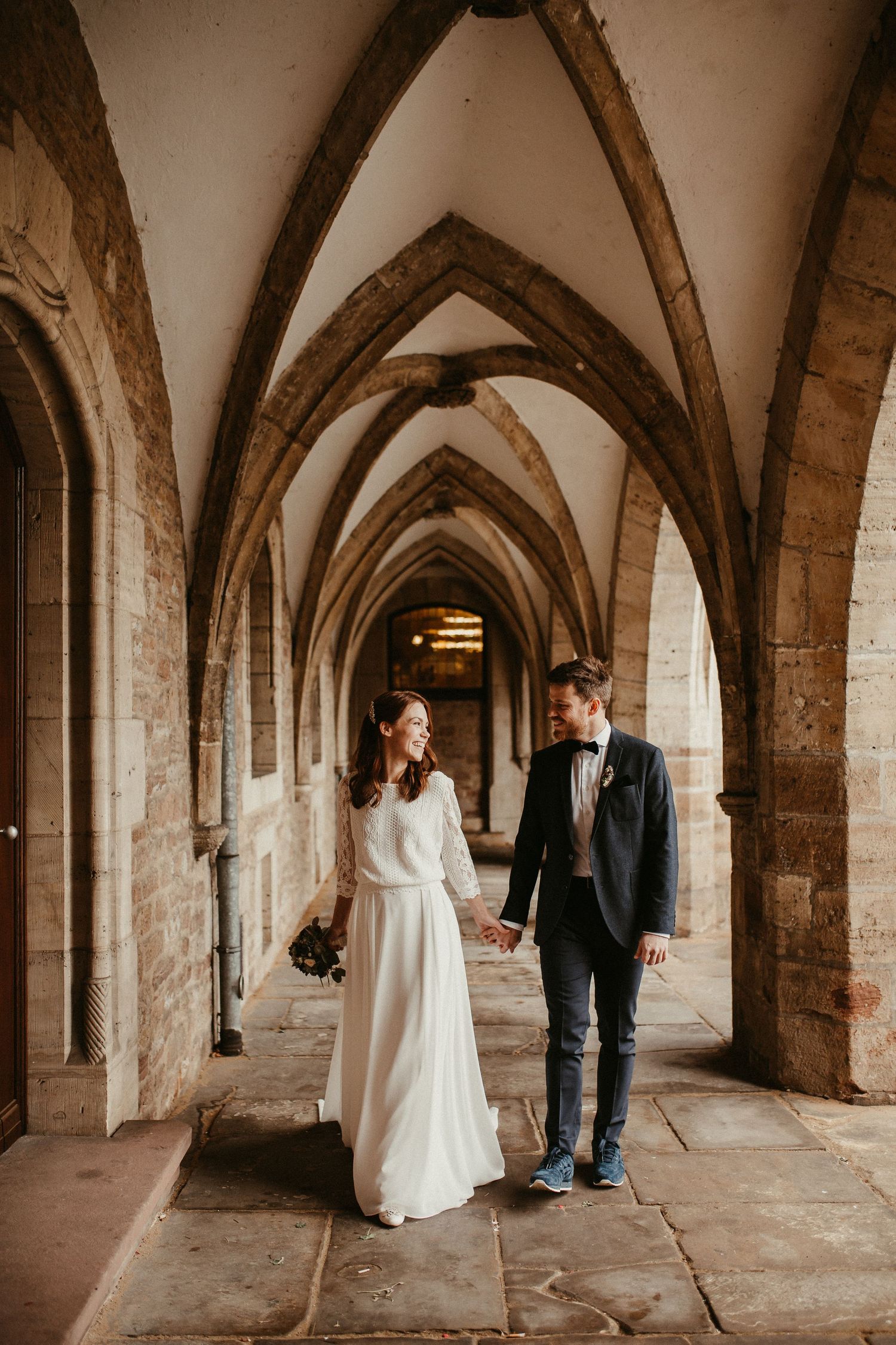 Paar geht in romantischer Hochzeitskleidung durch gotische Steinbögen.