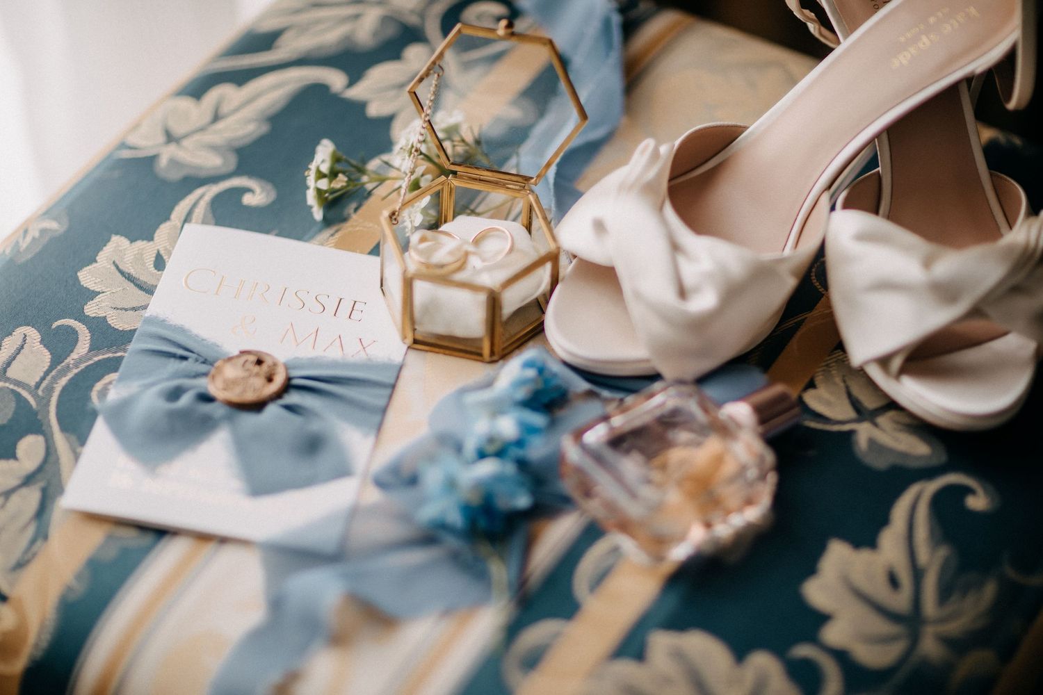 Hochzeitsdetails einschließlich weißer Satinschuhe, Einladung, Parfum und Ringen auf Blumenstoff.