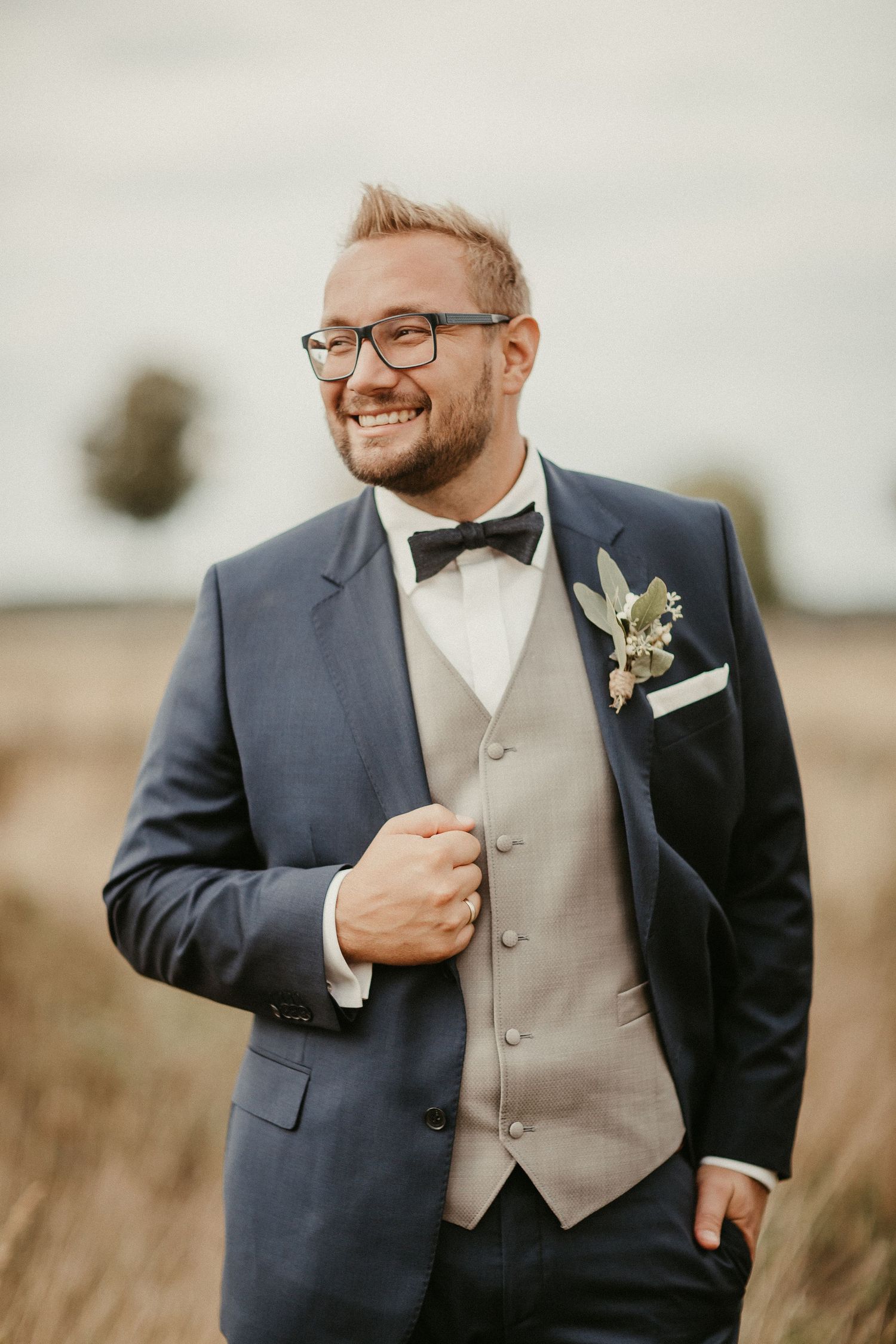 Bräutigam in marineblauem Anzug und Brille lächelt, während er Boutonniere vor natürlichem Hintergrund trägt.