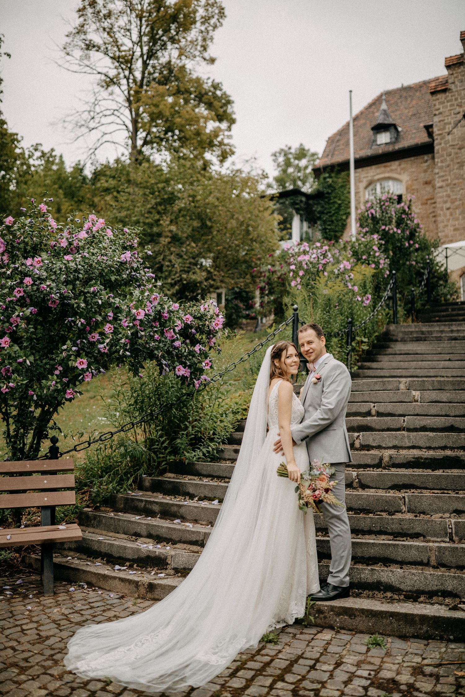 Romantisches Hochzeitsporträt auf Steinstufen mit blühendem Garten und historischem Gebäudehintergrund.