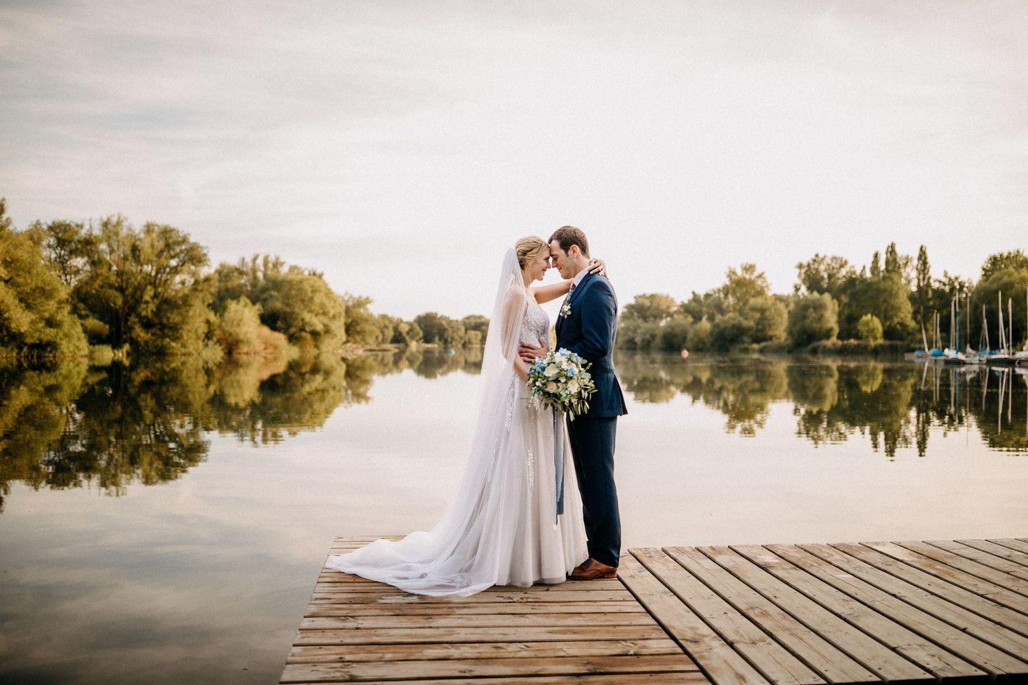 Hochzeitspaar umarmt sich auf einem Holzdock mit Blick auf einen ruhigen See bei Sonnenuntergang in Braunschweig.