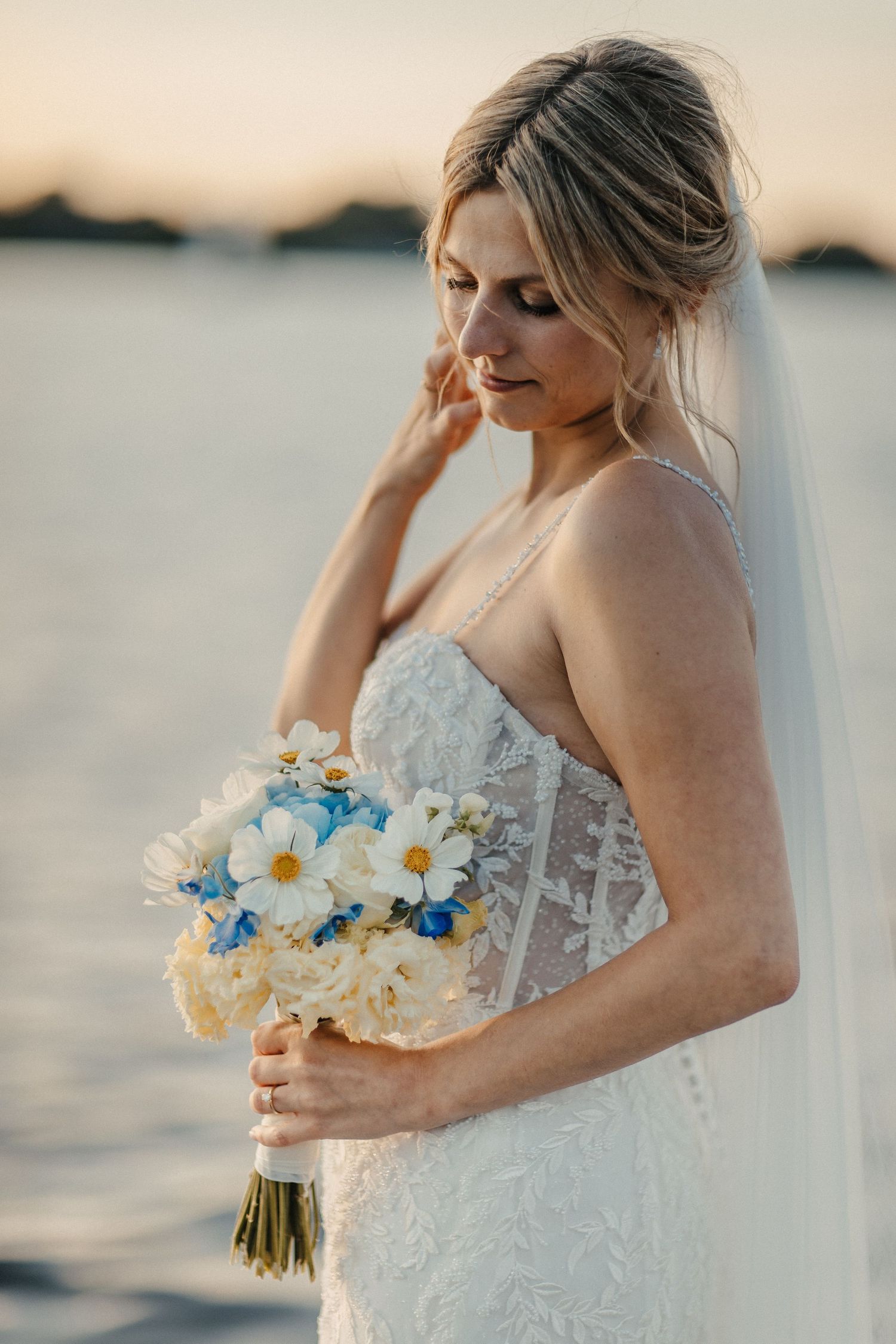Braut im Spitzenkleid mit Gänseblümchenstrauß während des Sonnenuntergangs-Strand-Fotoshootings.