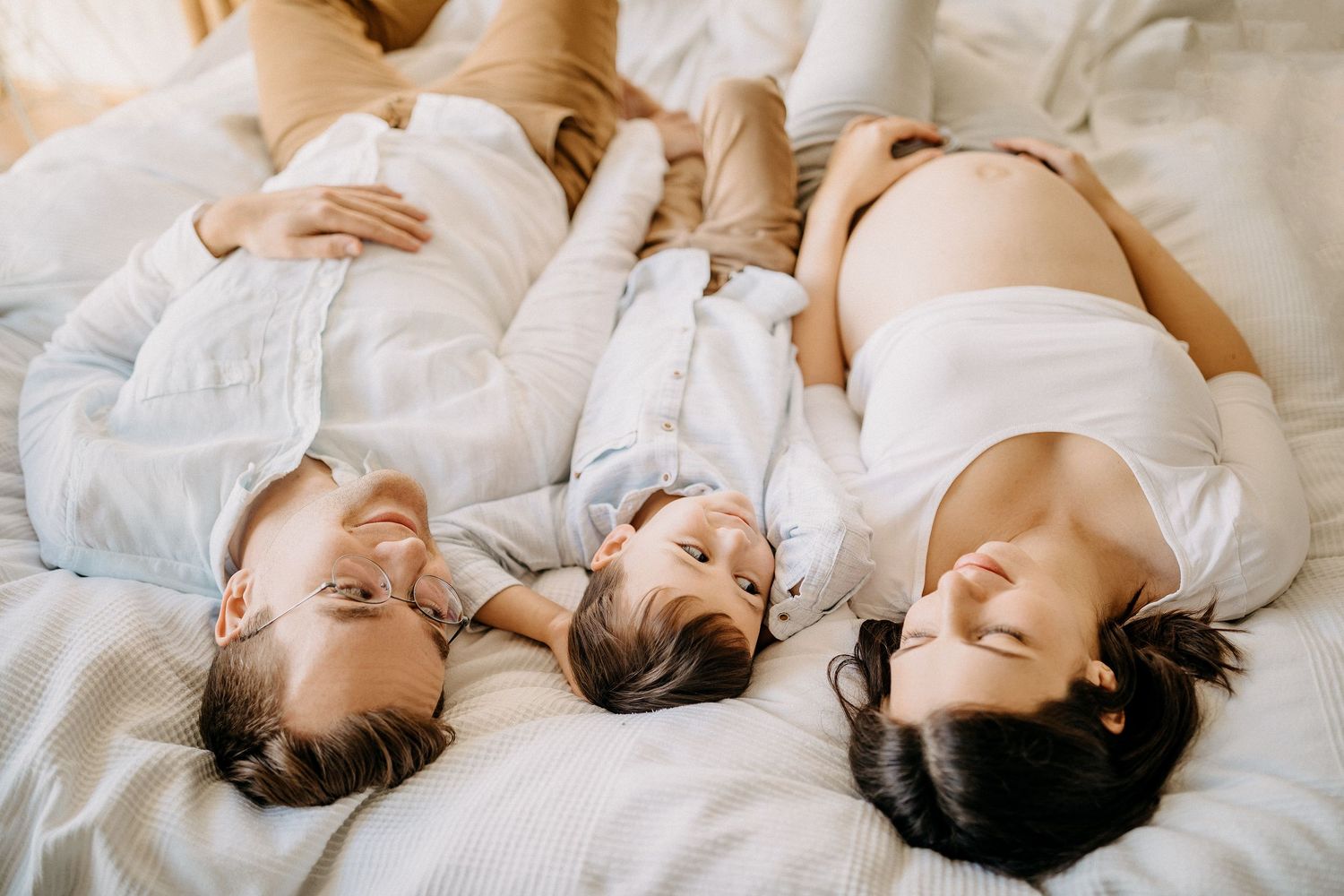 Familie liegt zusammen auf weißer Bettwäsche während eines intimen Lifestyle-Fotoshootings.