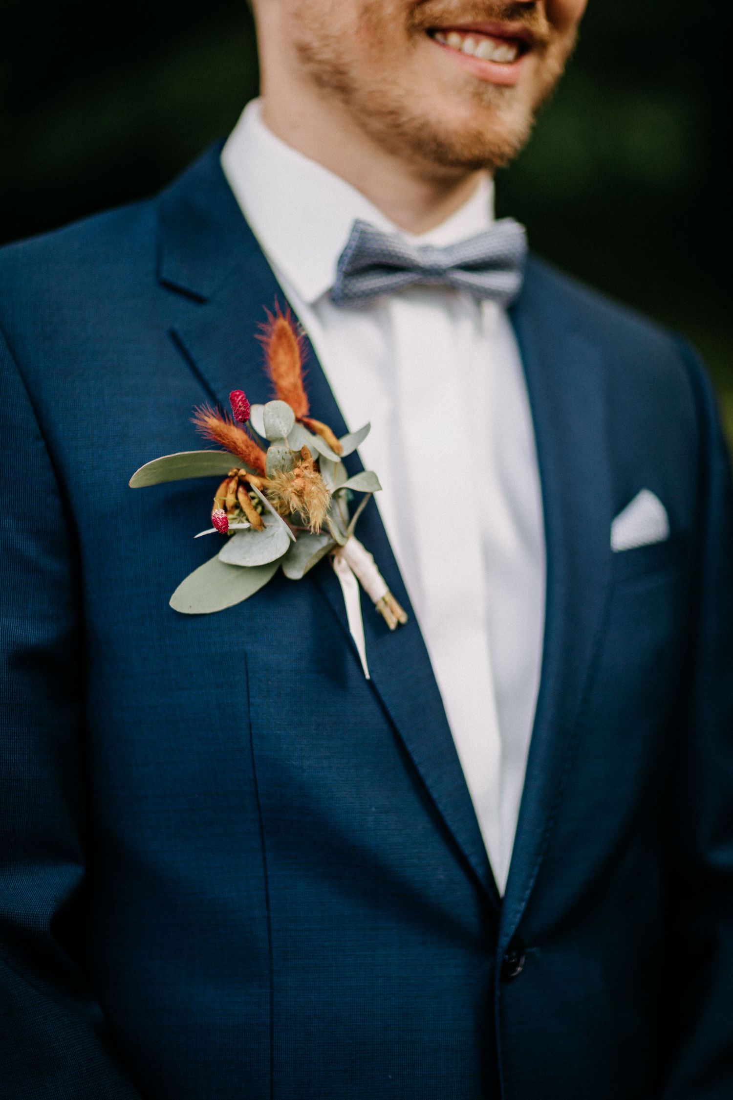 Nahaufnahme des marineblauen Anzugs Revers des Bräutigams mit orangefarbener und Eukalyptus-Boutonniere.