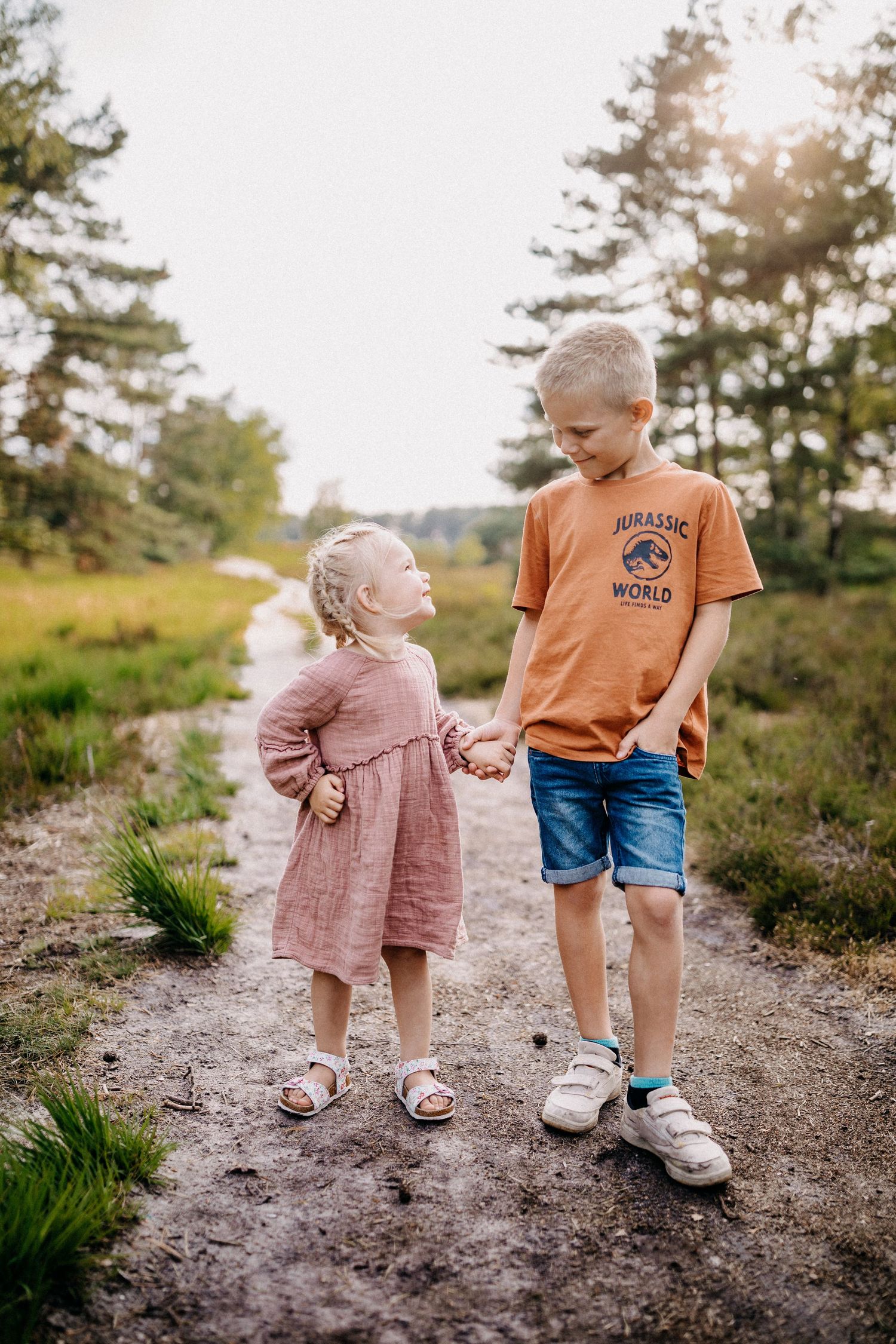 Zwei Kinder halten sich an den Händen, während sie auf einem unbefestigten Weg mit Kiefern im Hintergrund gehen in Gifhorn.