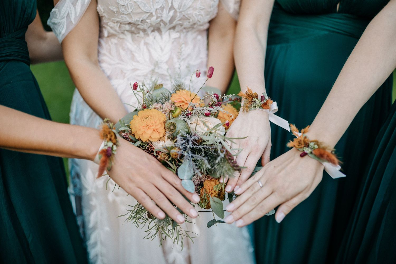 Mitglieder der Hochzeitsfeier zeigen florale Handgelenkskorsagen mit orangefarbenen Rosen vor smaragdgrünen Kleidern.