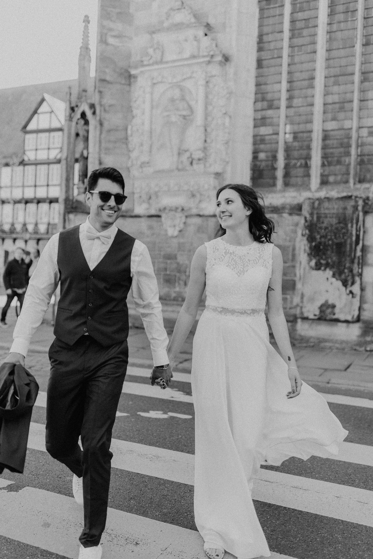 Schwarz-Weiß-Foto eines frisch verheirateten Paares, das über eine Straße vor einer gotischen Kirche geht.