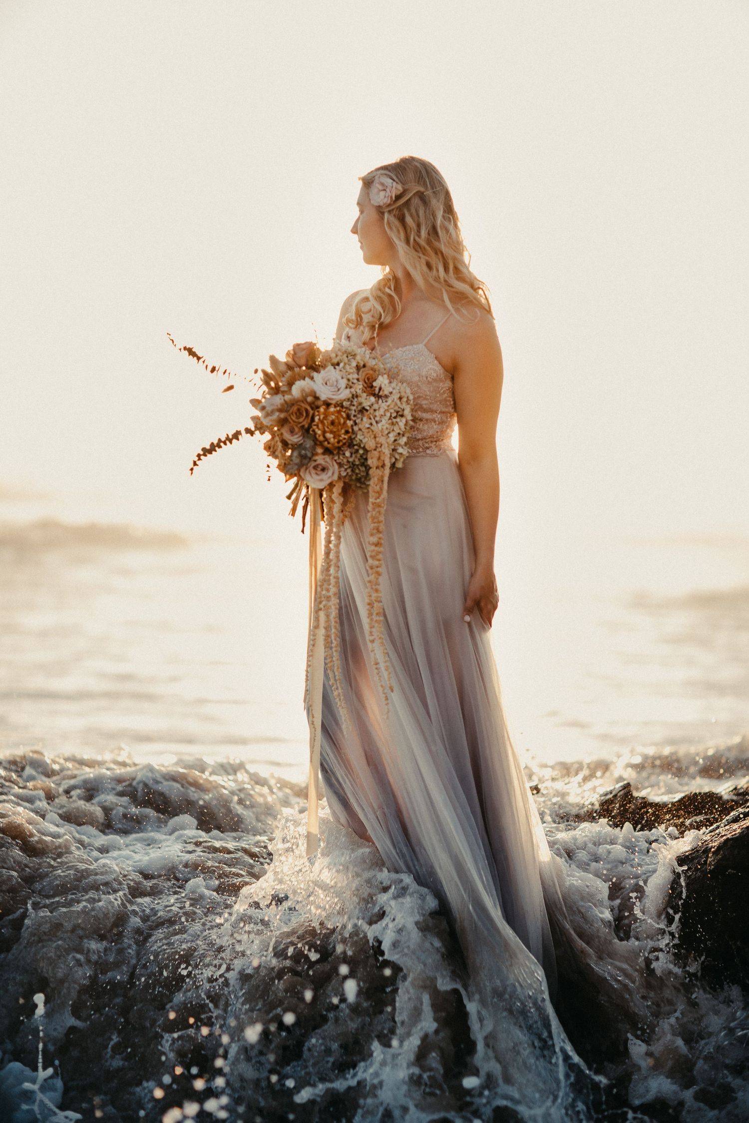 Ätherische Braut in fließendem grauem Kleid hält getrockneten Blumenstrauß, während sie in den Meereswellen steht in Südafrika.