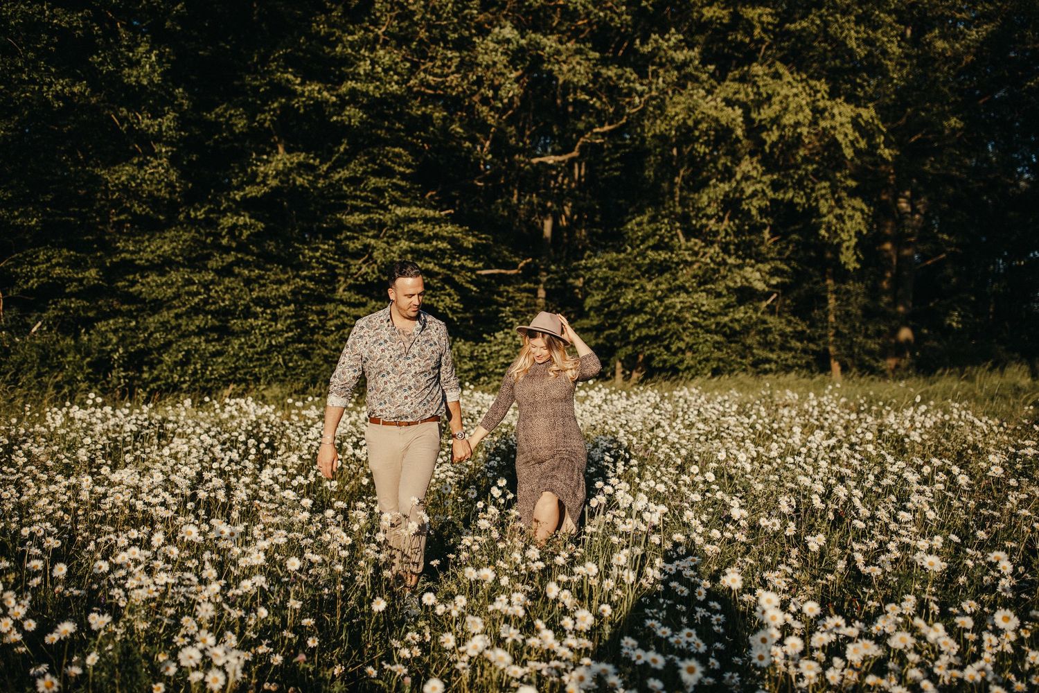 Paar geht Hand in Hand mit Babybauch durch eine blühende Wildblumenwiese.