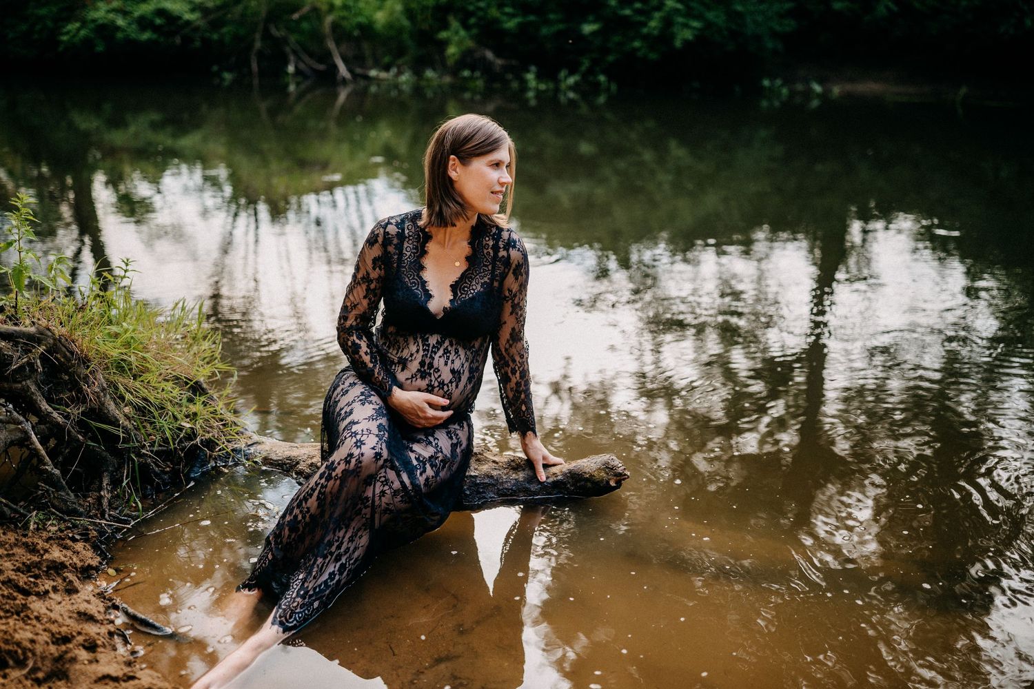 Schwangere in einem schwarzen Spitzenkleid posiert künstlerisch an einem reflektierenden Teich in einer stimmungsvollen Umgebung im Freien.