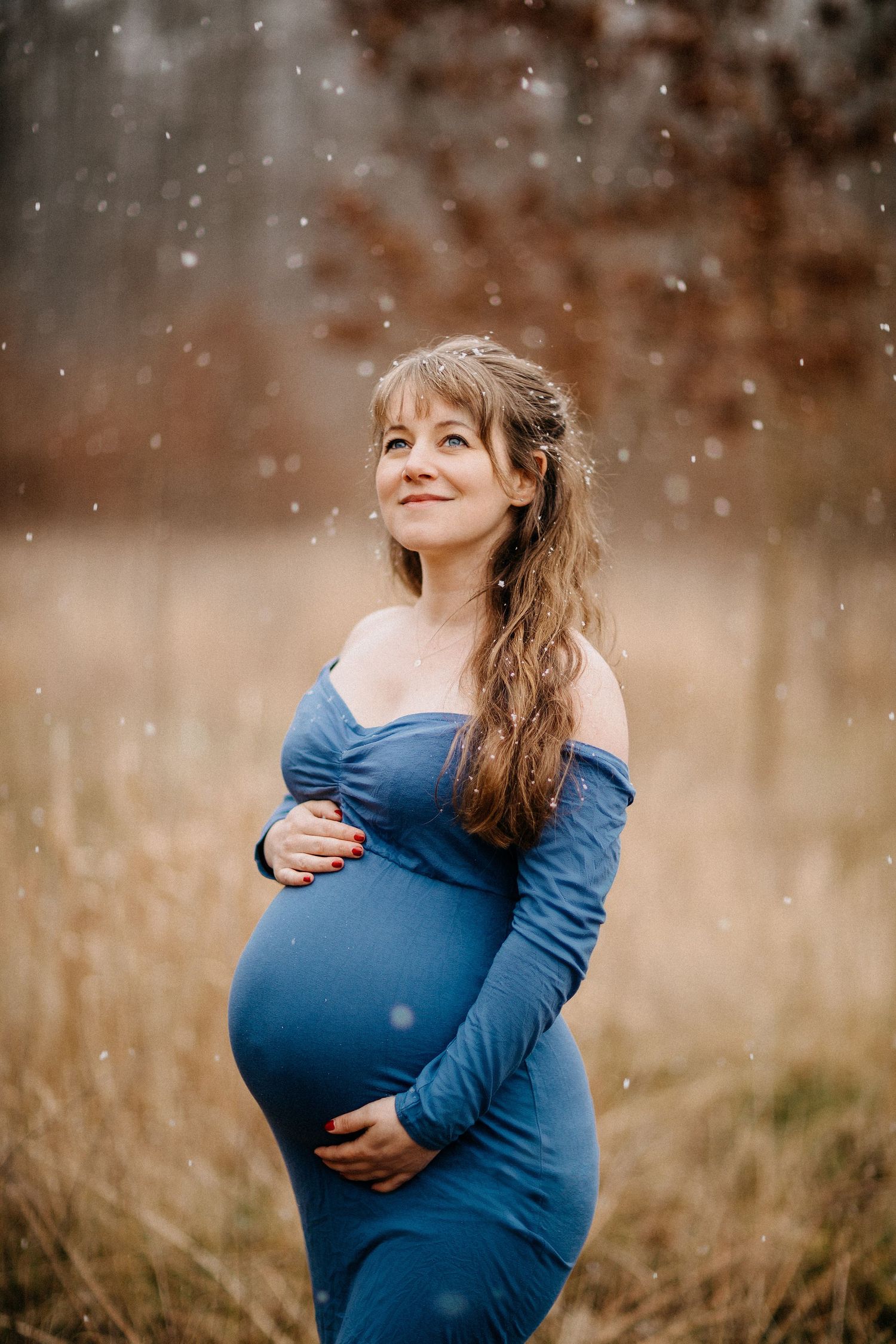 Schwangere Frau im blauen Kleid lächelt während des verschneiten Mutterschaftsfotoshootings.