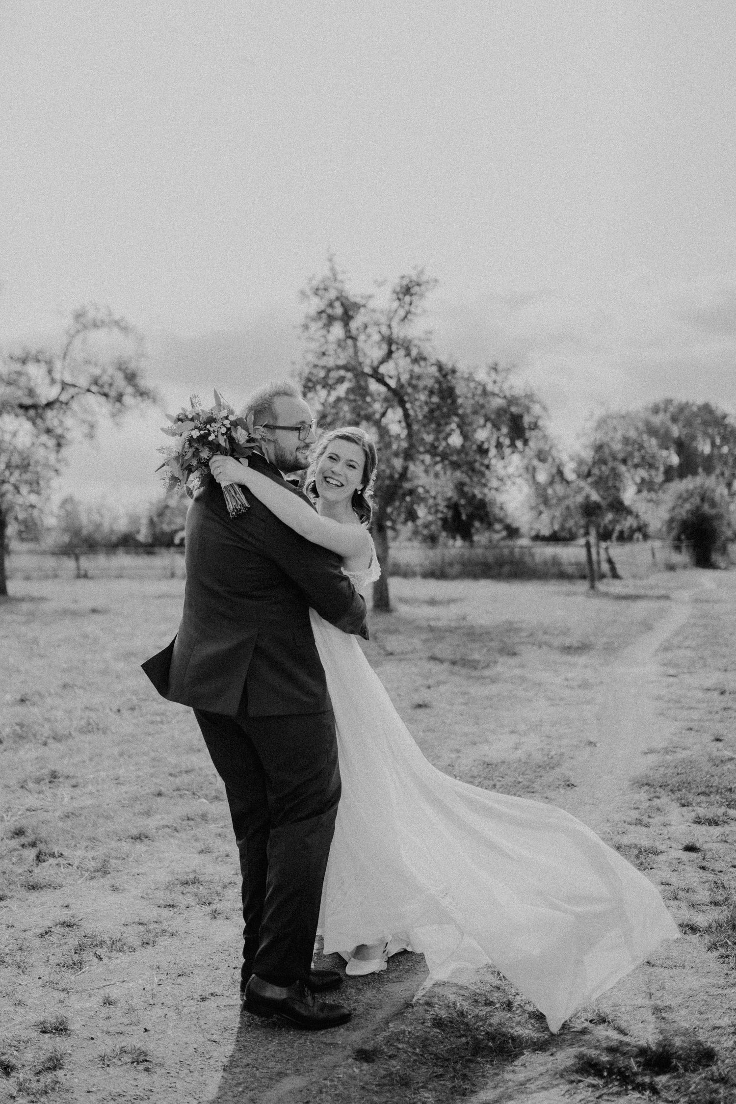 Schwarz-Weiß-Foto eines Paares, das zusammen auf dem Feld mit fließendem Hochzeitskleid tanzt.