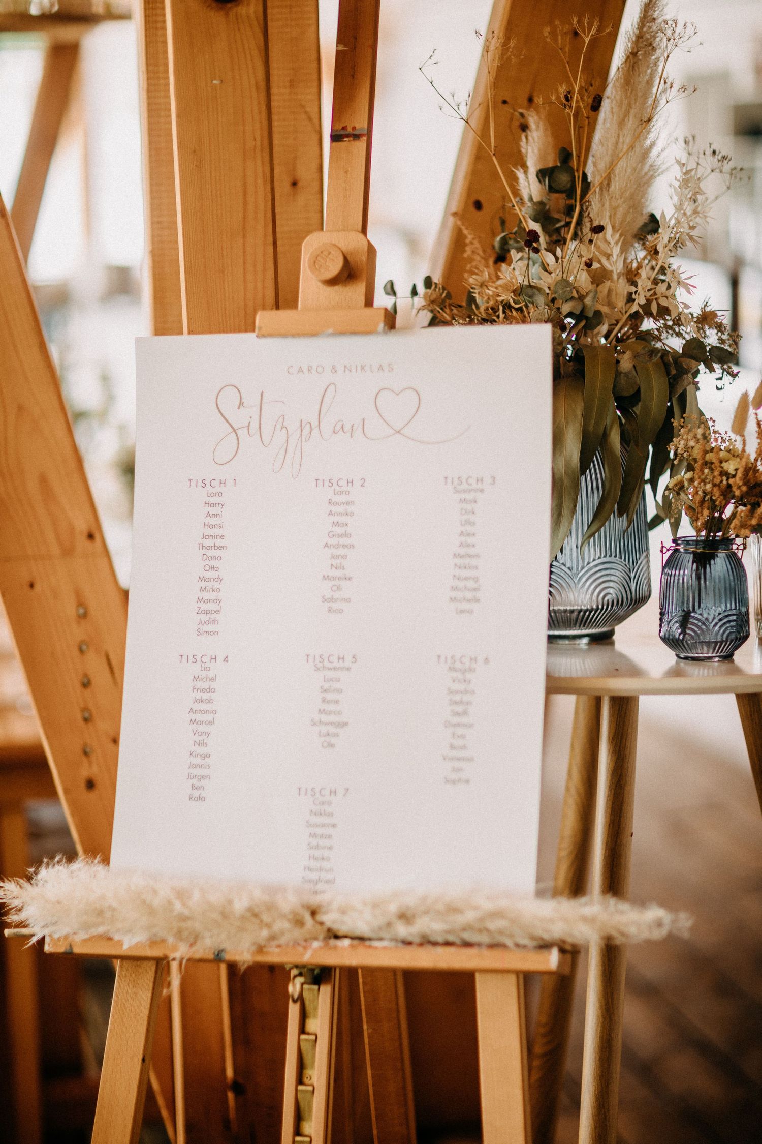 Hochzeitssitzplan auf einer Holzstaffelei mit dekorativen getrockneten Blumen in blauen Vasen.