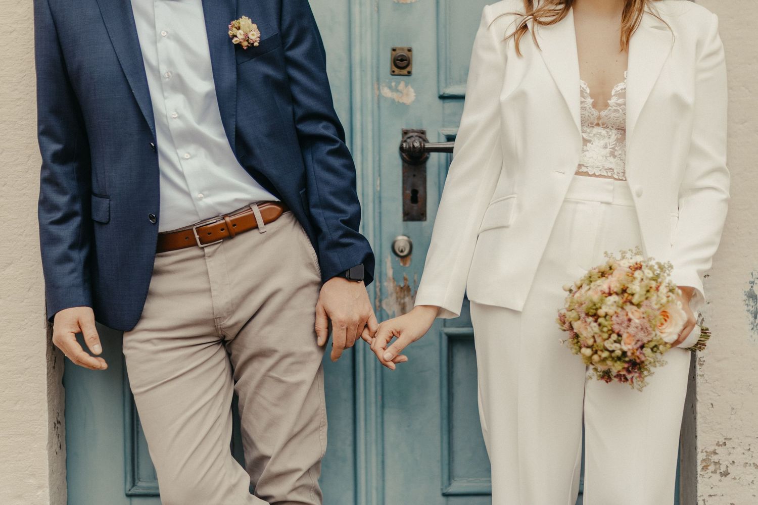 Modernes Paar in Anzug und weißem Blazer hält Händchen gegen die blaue Tür.