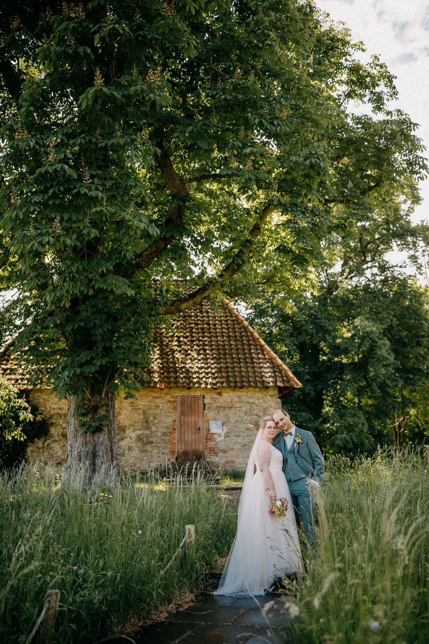 Ein romantisches Hochzeitsporträt in der Nähe eines rustikalen Steingebäudes, umgeben von üppig grünen Bäumen.
