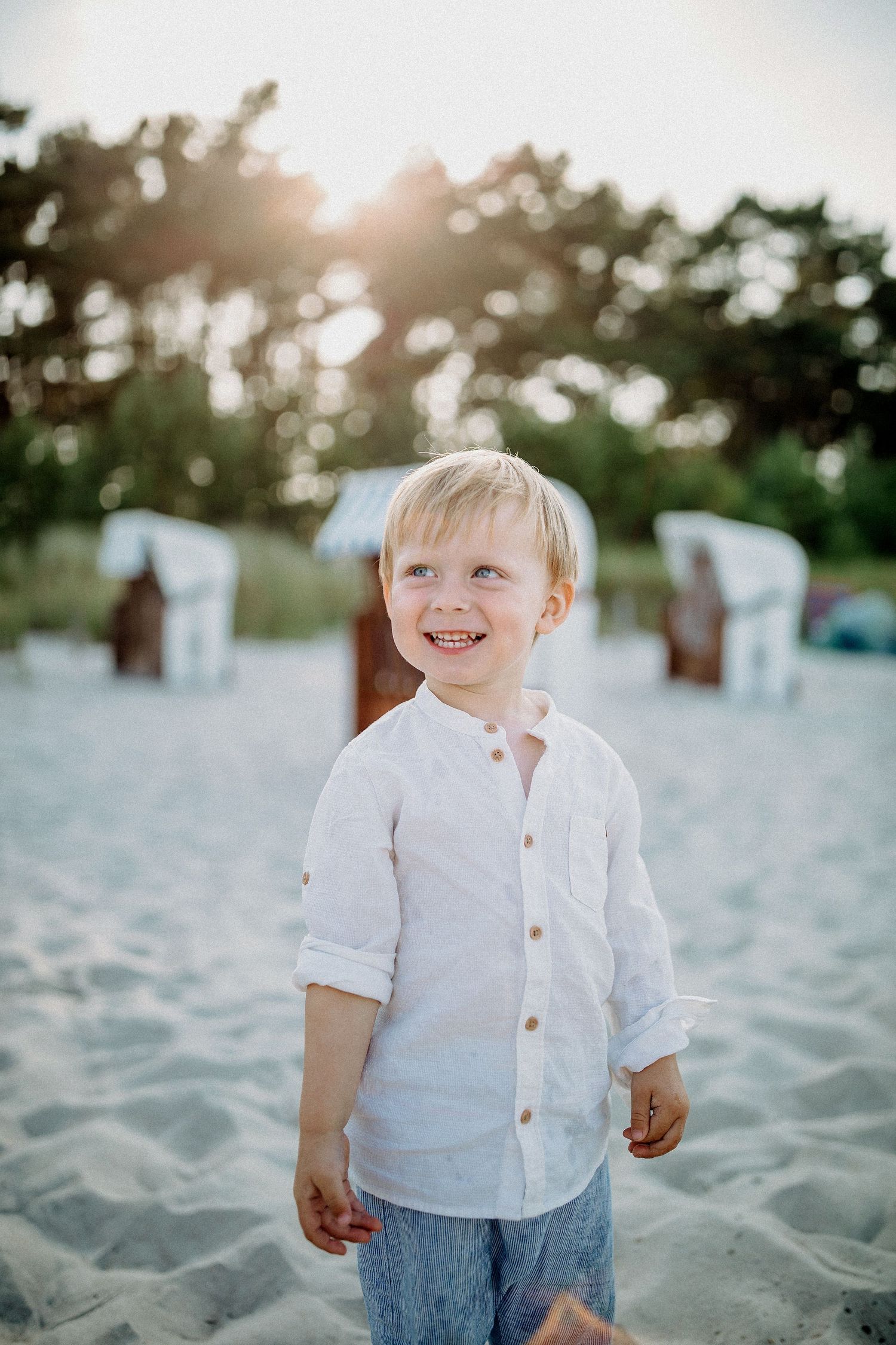 Junge im weißen Hemd lächelt am Sandstrand mit Liegestühlen im Hintergrund.