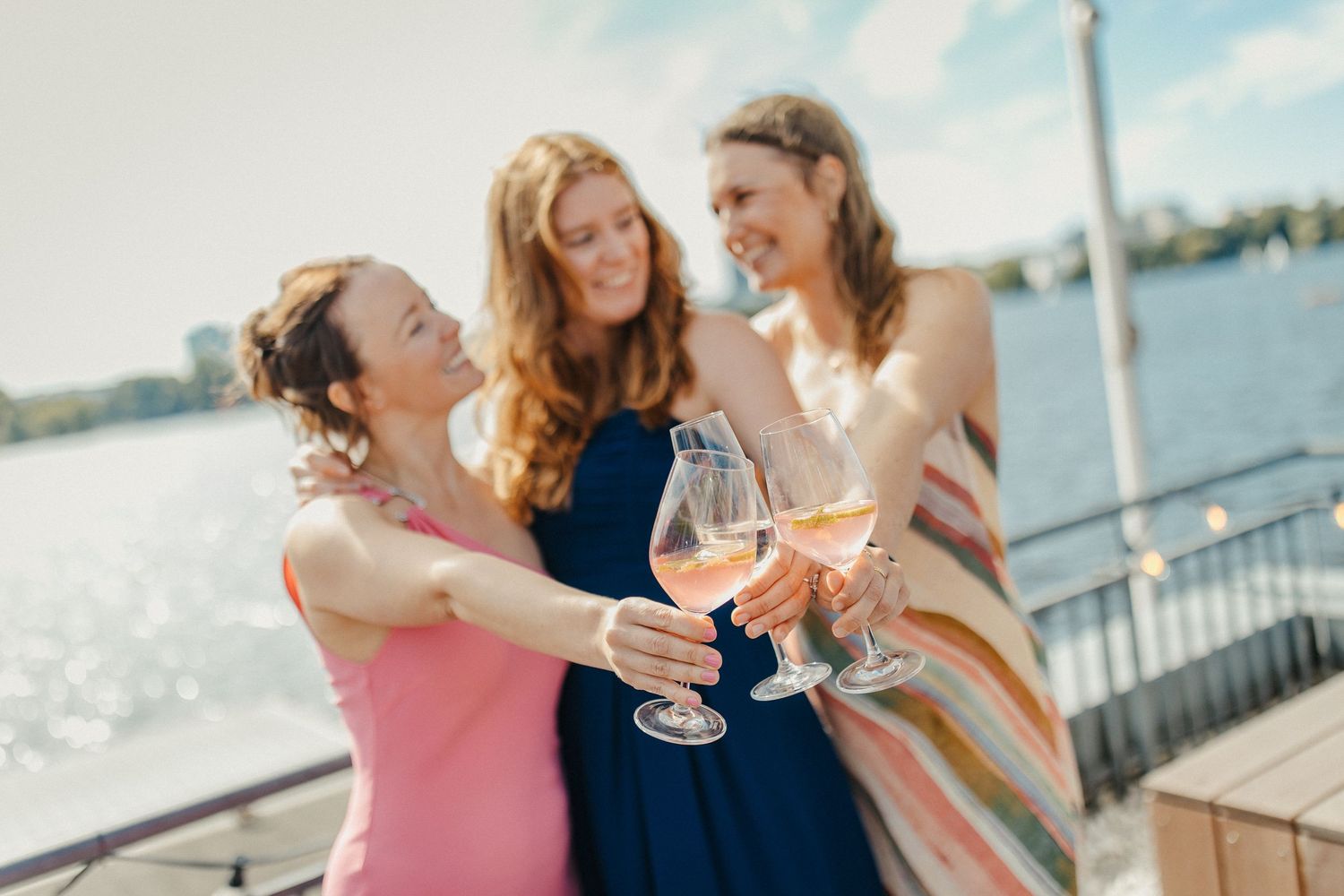 Drei Freunde in bunten Kleidern stoßen mit Weingläsern auf einer sonnigen Terrasse am Wasser an.