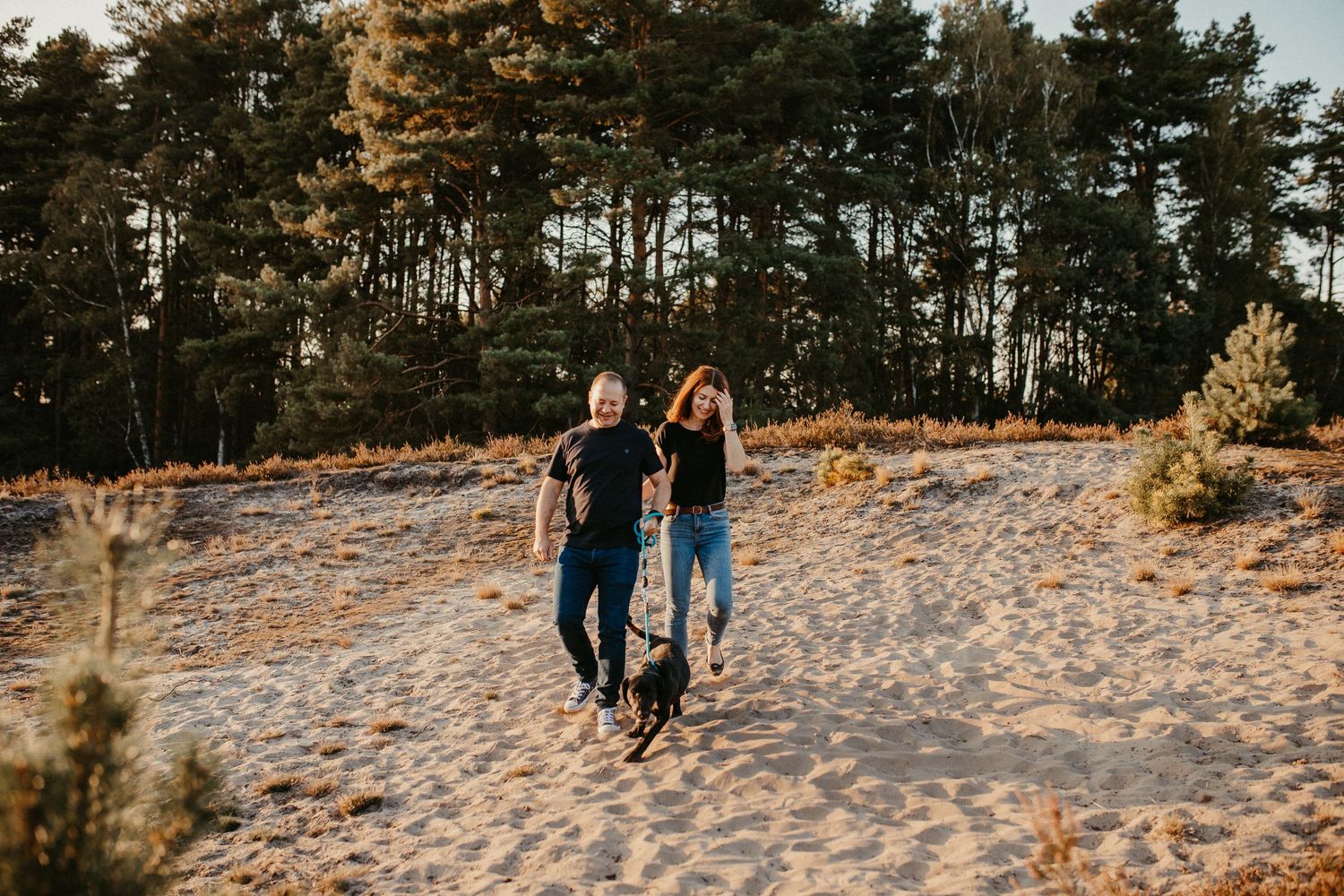 Ein Paar geht Hand in Hand auf einem Sandstrandweg bei Sonnenuntergang mit Kiefern im Hintergrund mit Hund.