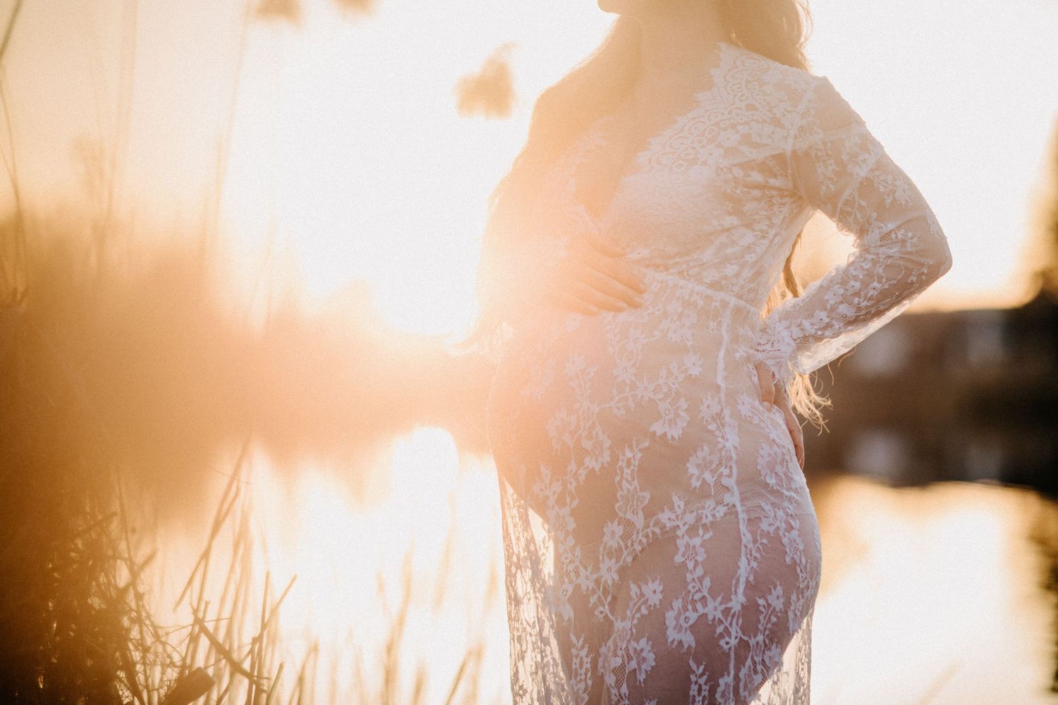 Silhouette einer schwangeren Frau in weißem Spitzenkleid vor goldenem Sonnenuntergangslicht.