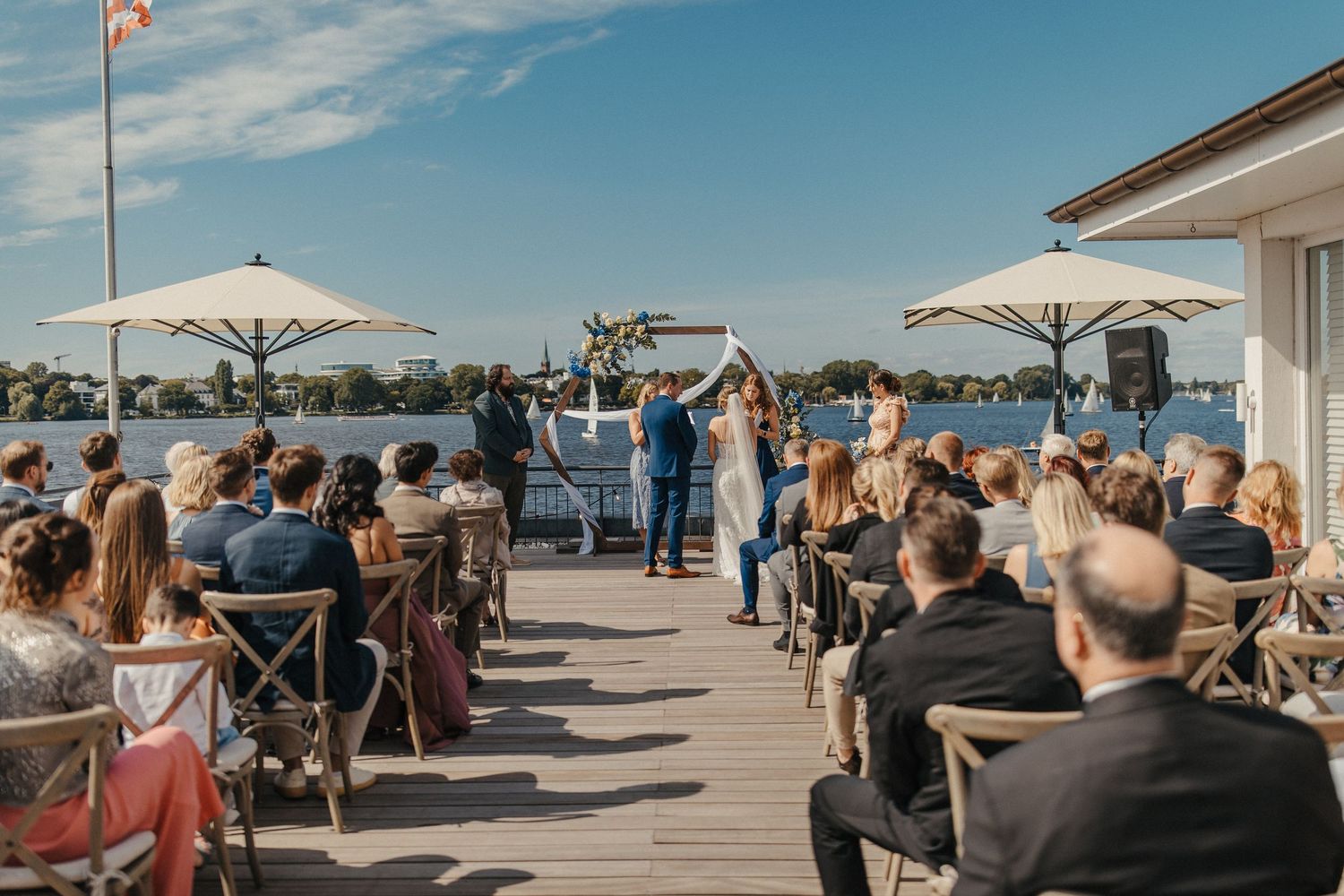 Eine Hochzeitszeremonie am Wasser mit weißen Sonnenschirmen und Gästen, die auf einer Terrasse sitzen.