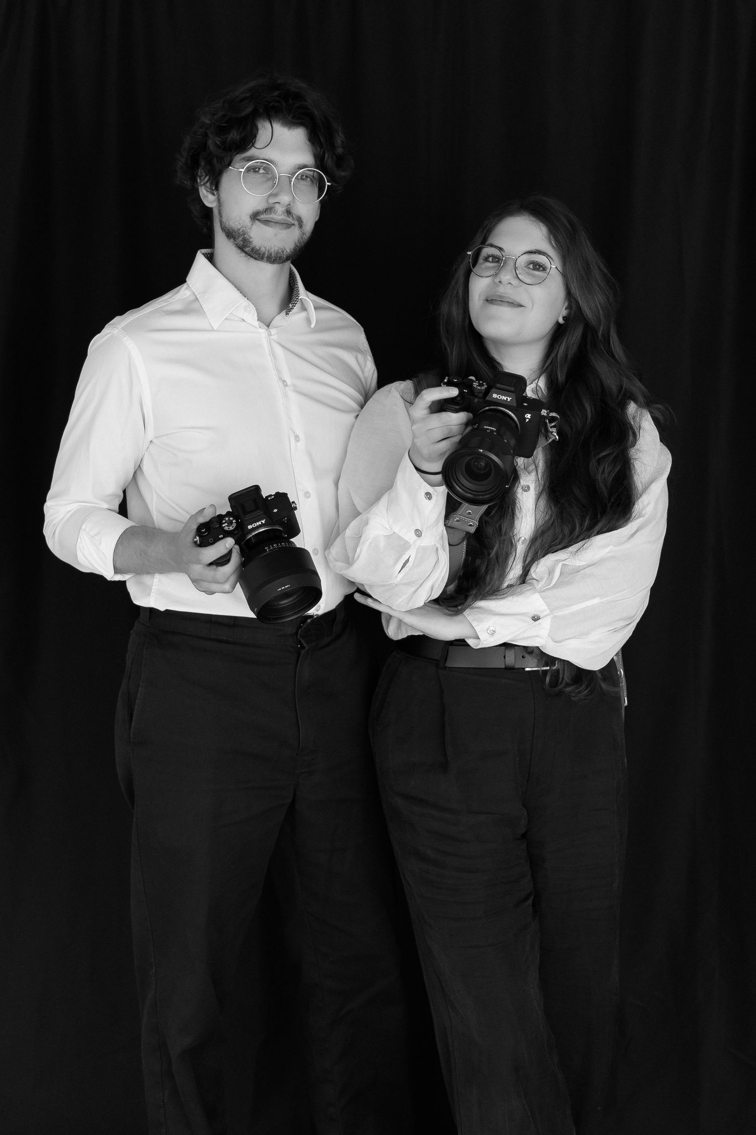 A black and white portrait of two photographers in matching white shirts holding professional cameras against a dark background.