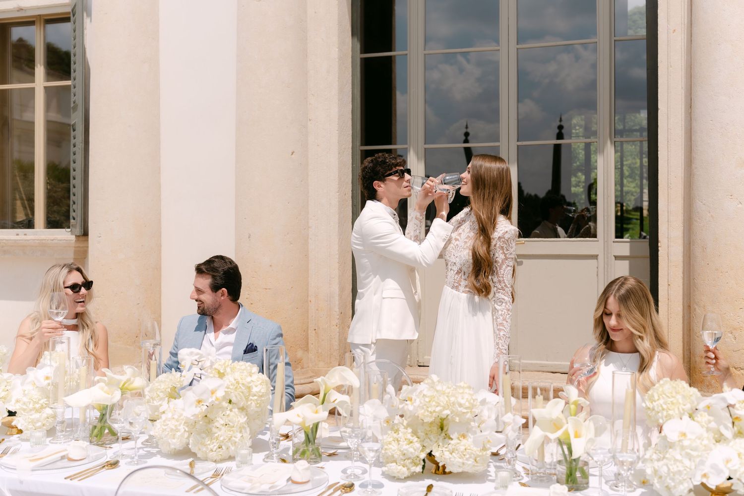 Elegant wedding reception table decorated with white flowers as guests celebrate during a toast at luxurious venue.
