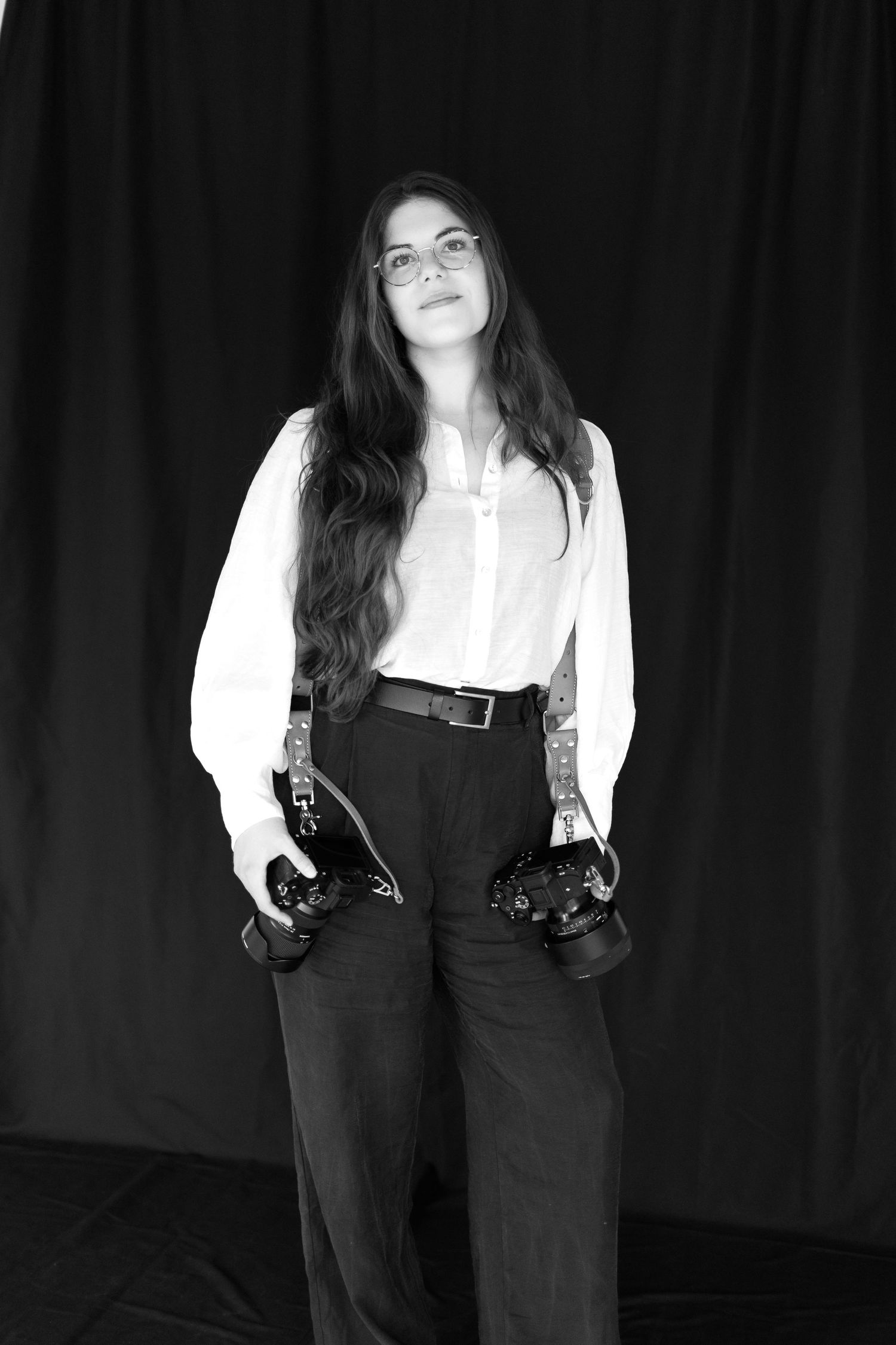 Black and white portrait of a photographer wearing glasses, white shirt and dark pants holding two cameras against dark backdrop.