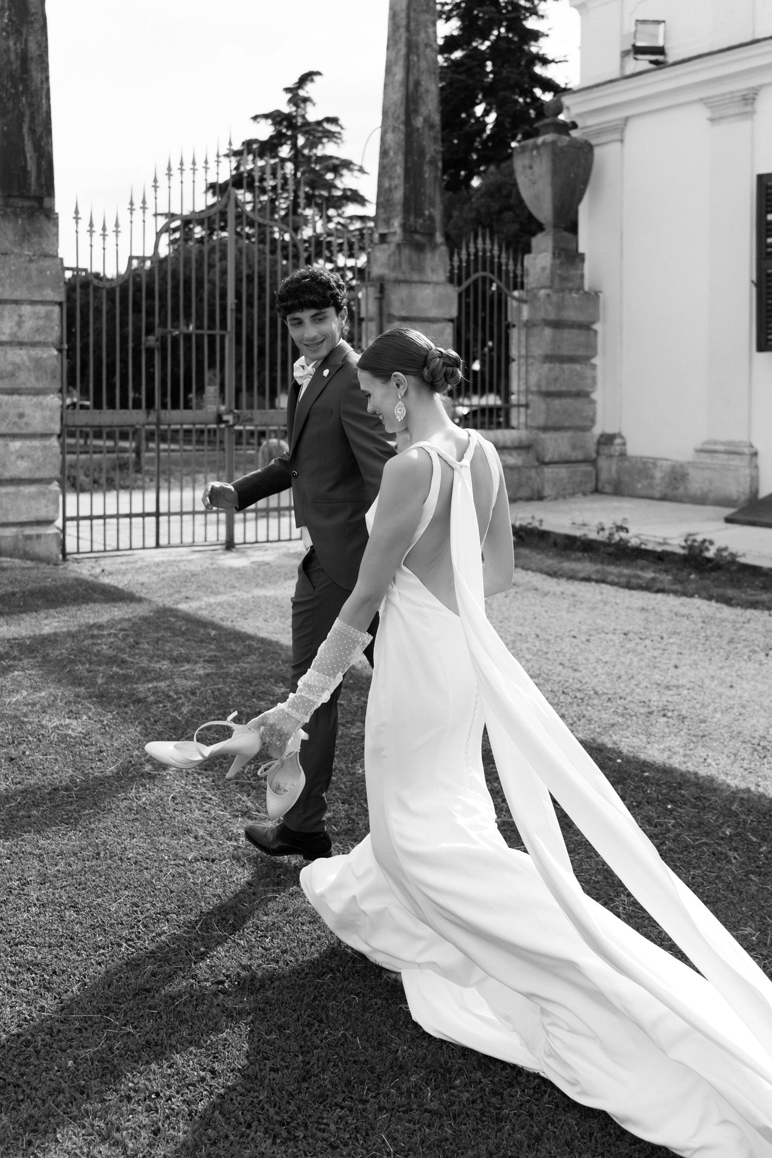 A couple in wedding attire walking together on garden grounds with an elegant iron gate and white building in background.