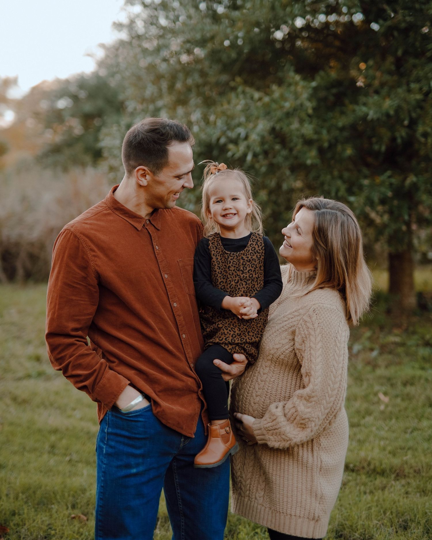A family of three shares a warm moment outdoors in autumn, wearing cozy sweaters while standing together in nature.