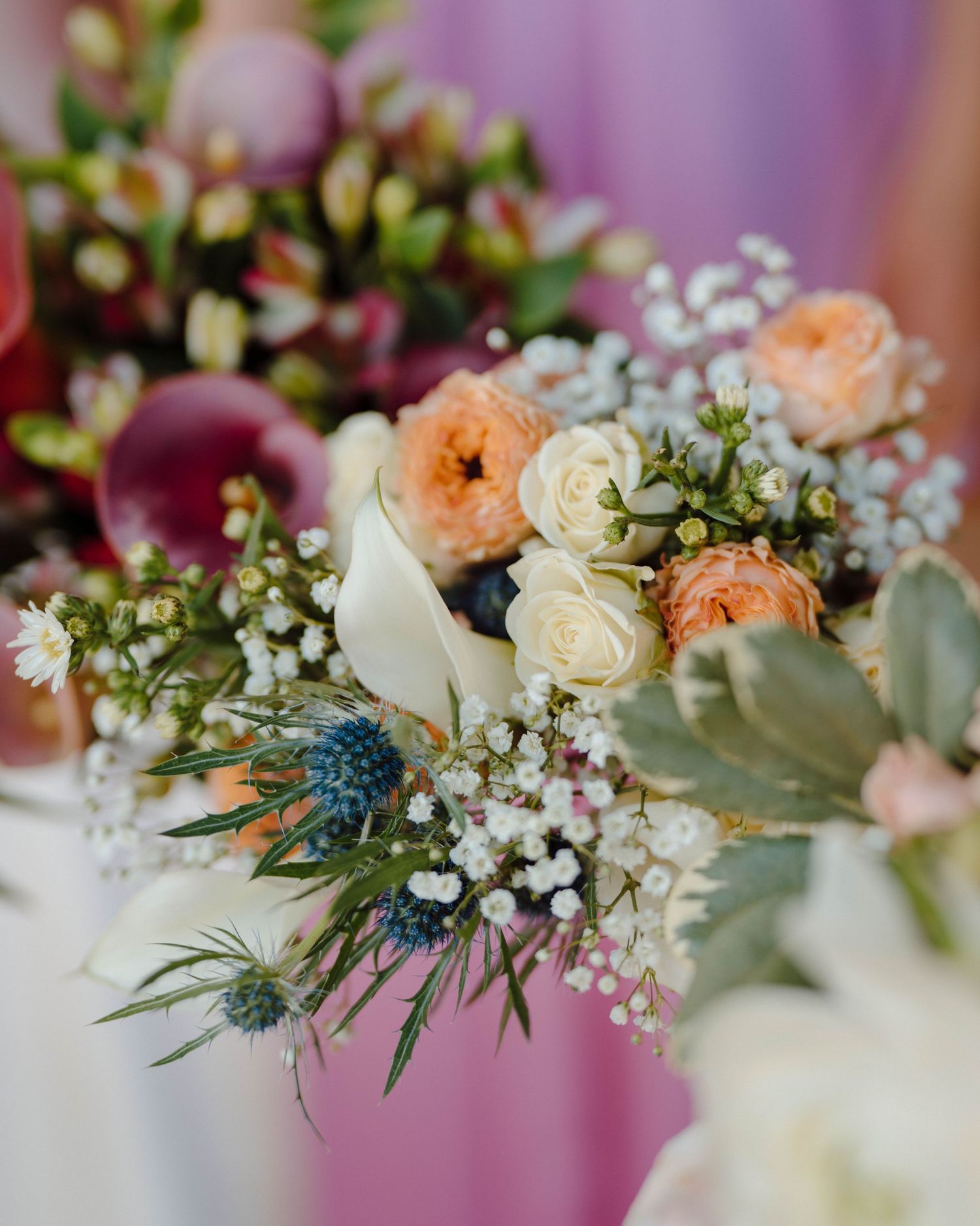 Wedding floral arrangement with peach roses, baby's breath, blue thistle, and sage greenery against a soft purple backdrop.