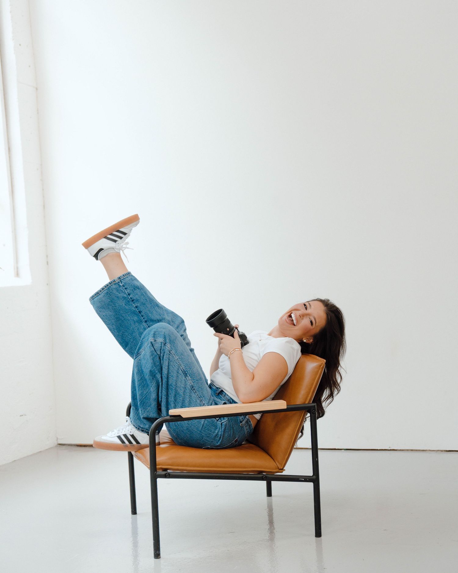 Person in casual denim and white top lounges playfully in a tan armchair while using a mobile device.