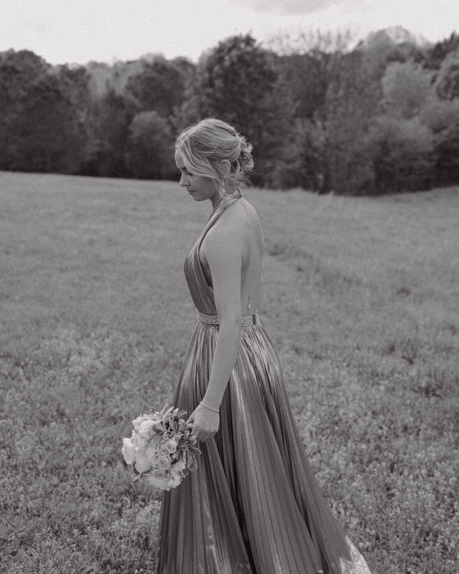 A person in an elegant pleated gown holds a floral bouquet while standing in a sunlit meadow, captured in black and white.
