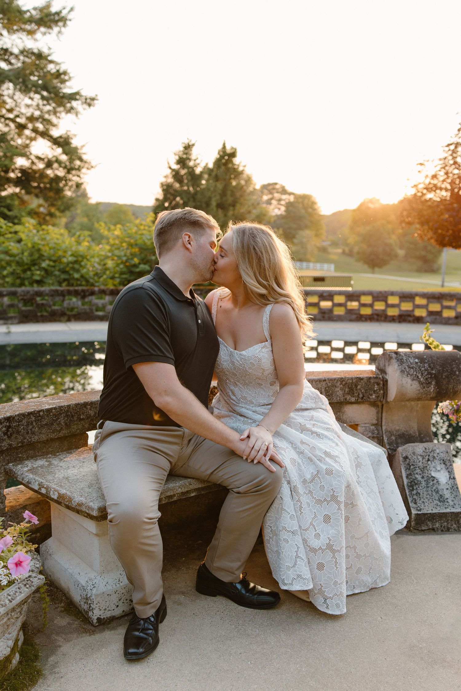 A couple shares a romantic moment at sunset on a stone bench in a garden setting with mountain views.