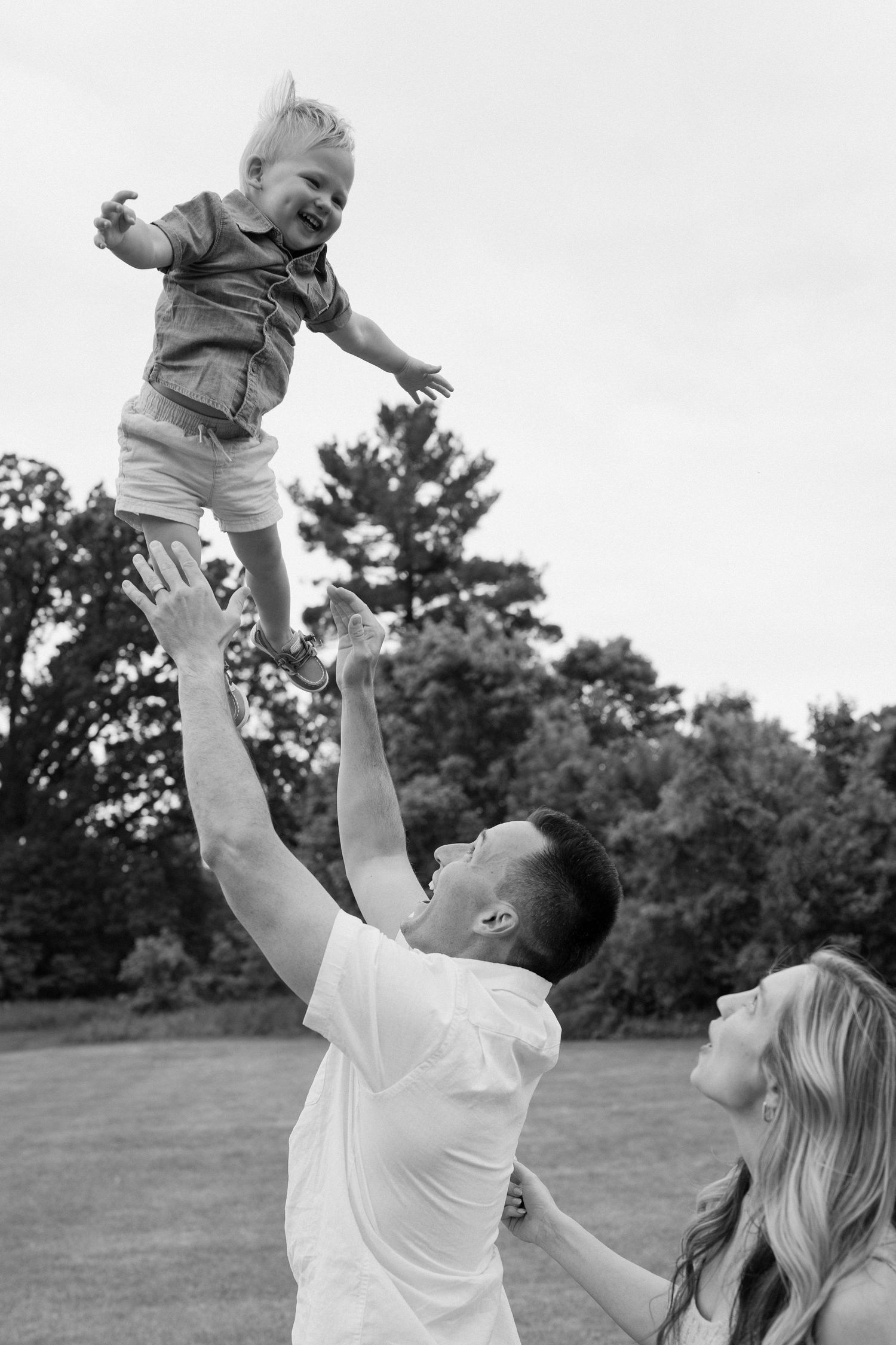 Person in white lifts and plays with small child in an open grassy field during a playful outdoor moment.